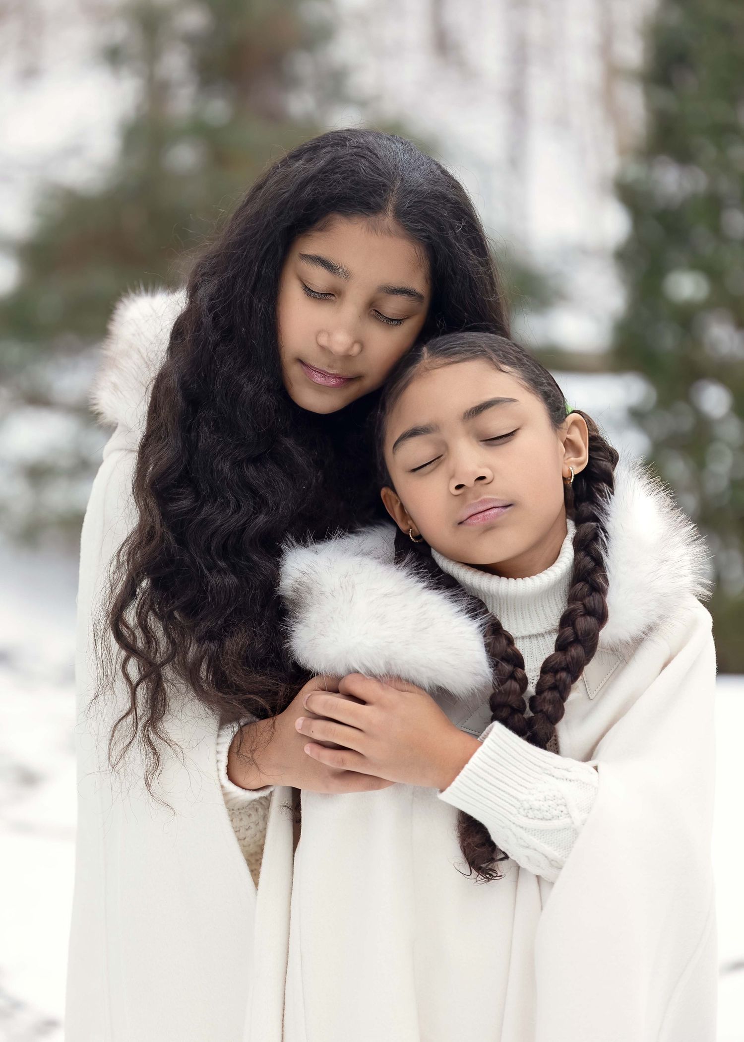 Two young girls in white winter coats embrace tenderly in a snowy outdoor setting with evergreen trees.