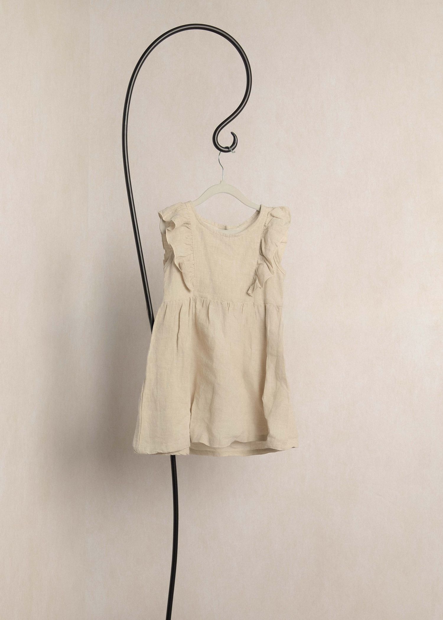 Cream linen children's dress with ruffle shoulders on a decorative hook hanger against a neutral wall.