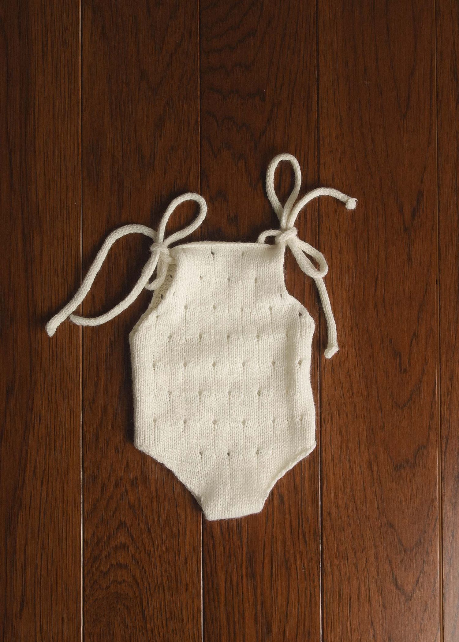 Cream knit baby romper with tie shoulders and eyelet detail, laid flat on wood floor.