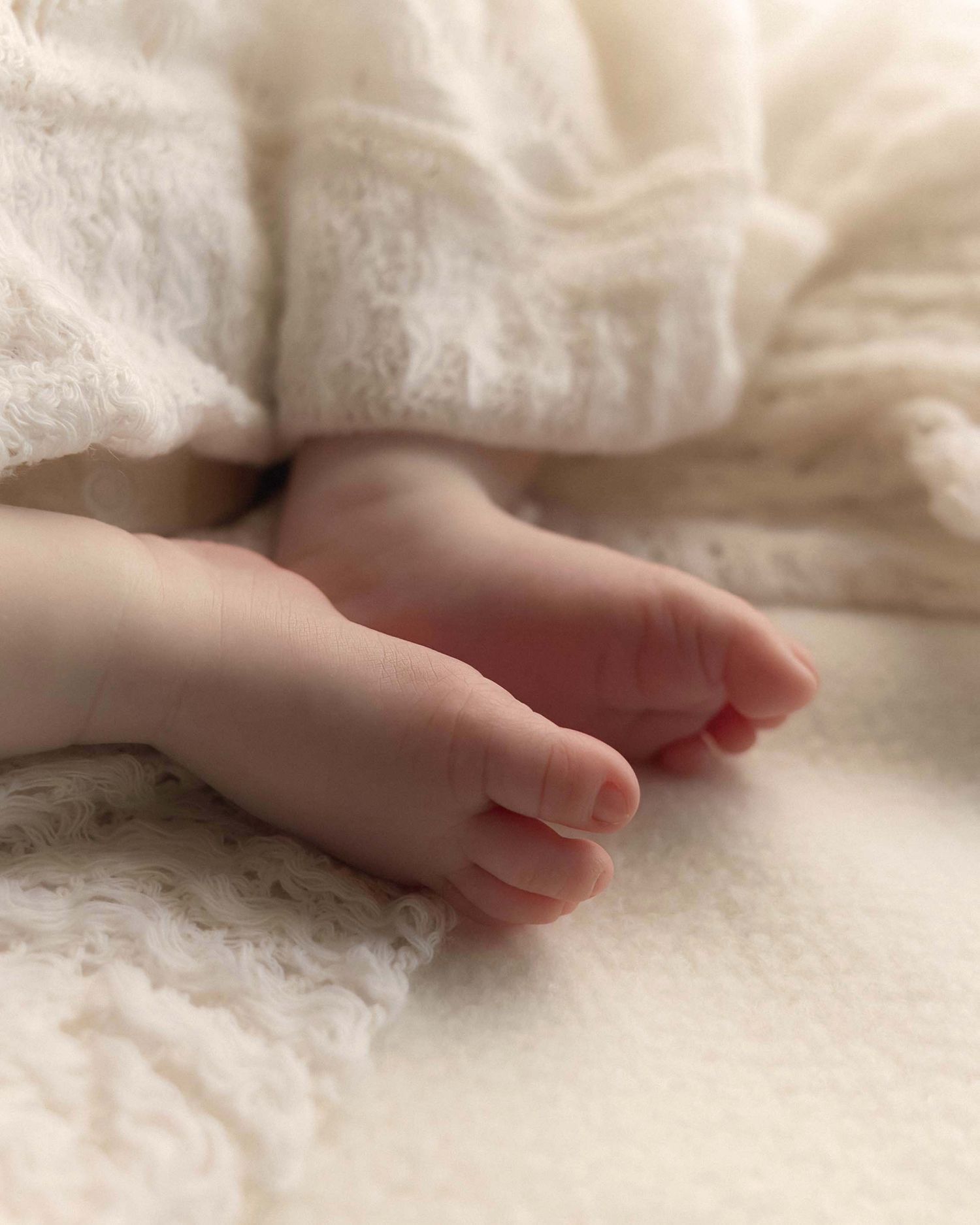 Close-up of tiny newborn baby feet resting on a soft white lace blanket in a delicate portrait photo.