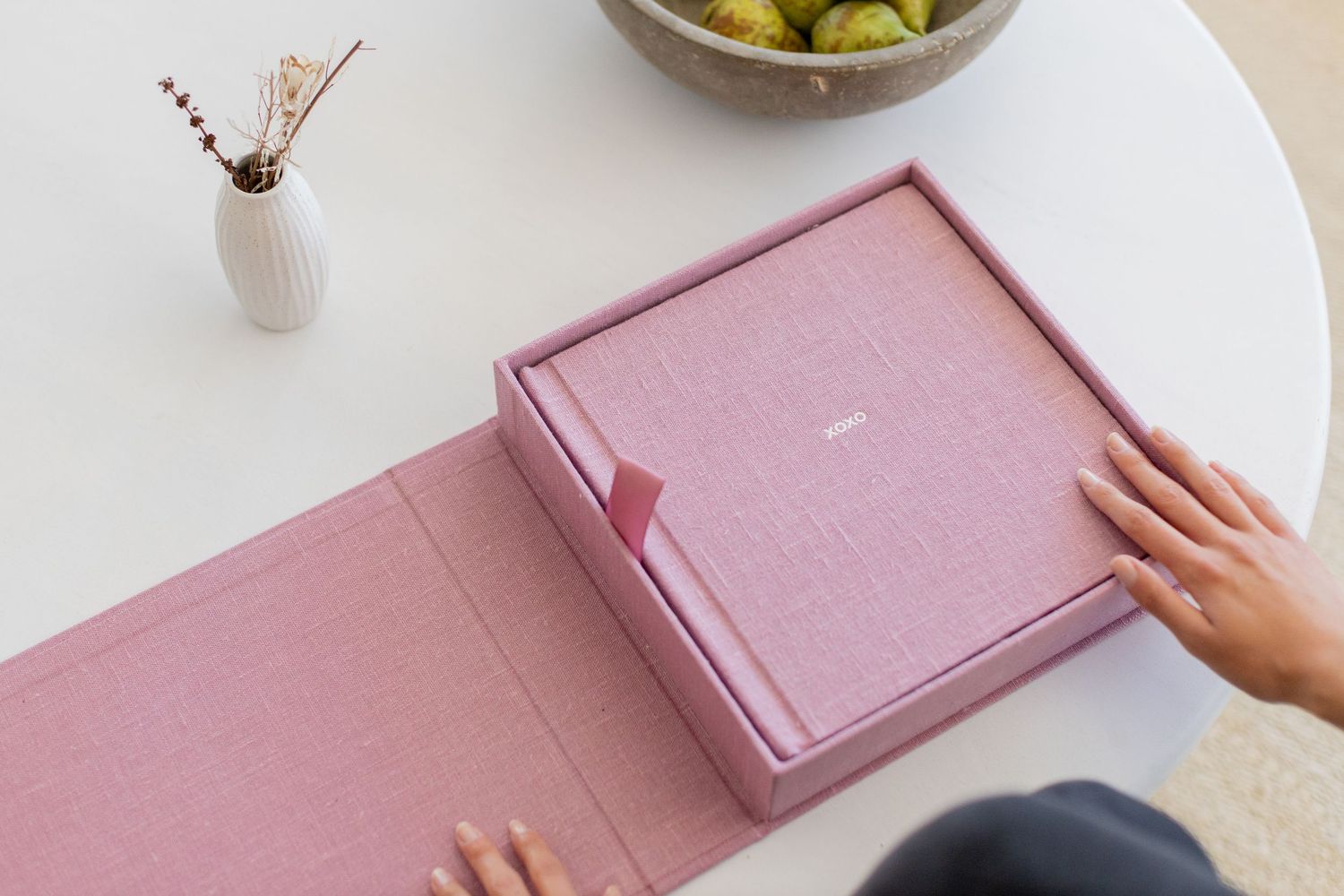 Hands opening a pink linen photo album box on a white table with a vase and bowl of fruit nearby.