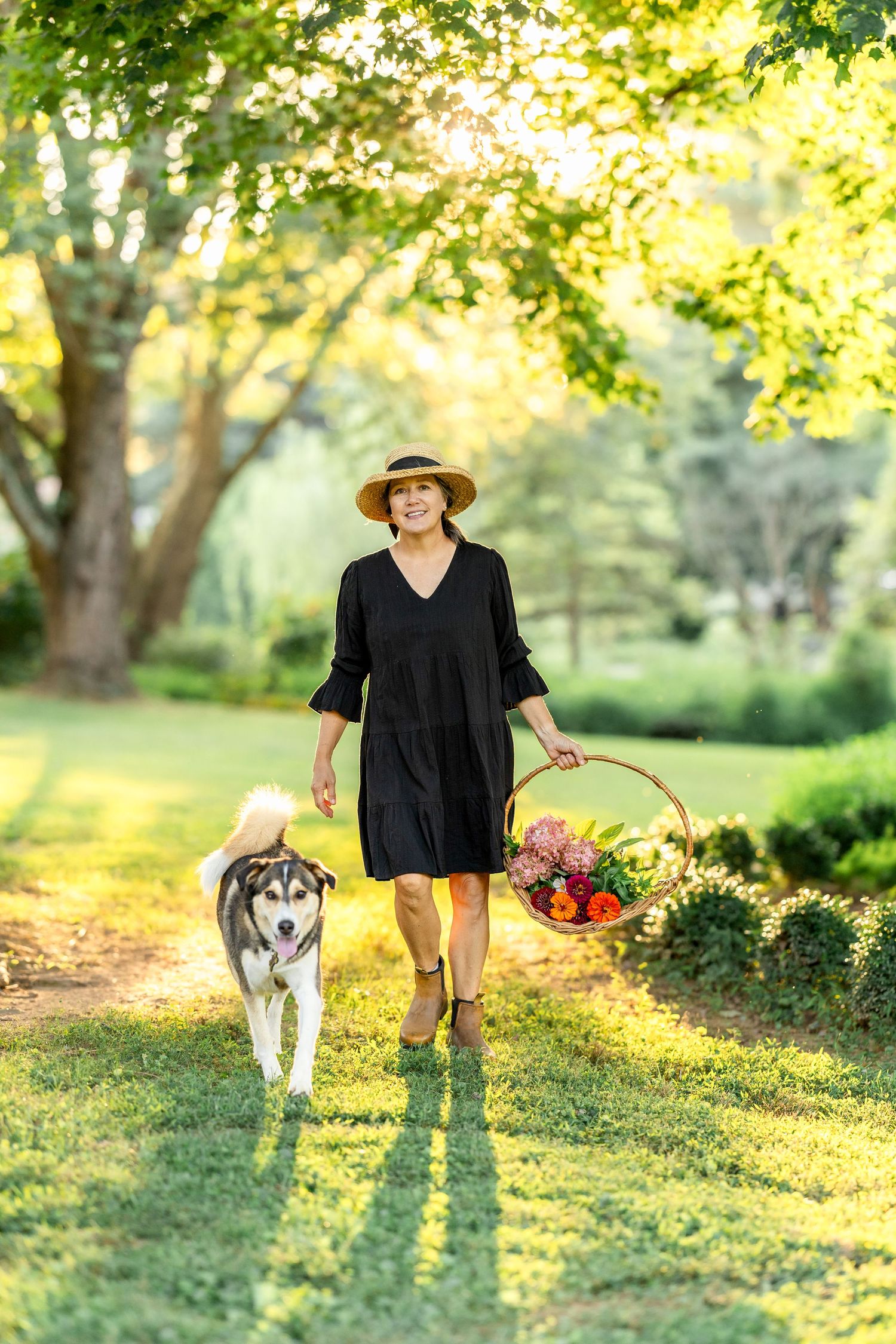 Person in black clothing walks with husky dog through sunlit park carrying flower basket.