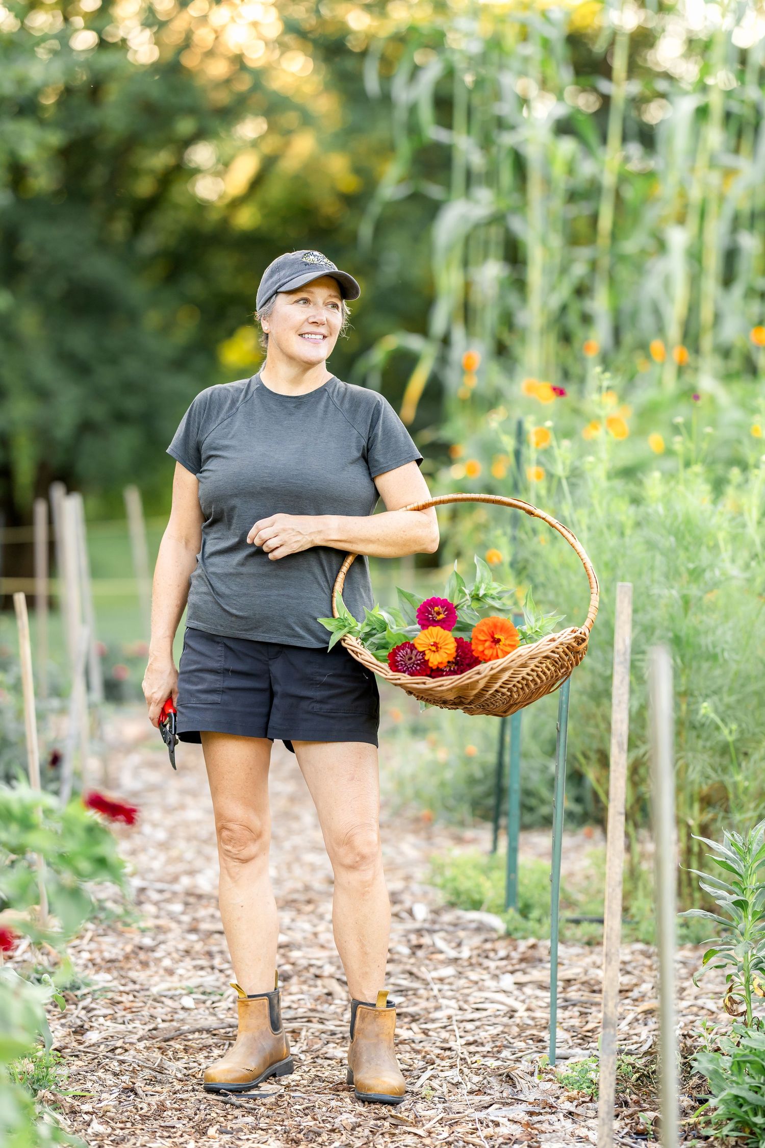 A wicker basket filled with fresh vegetables and flowers sits on a mulched garden path.