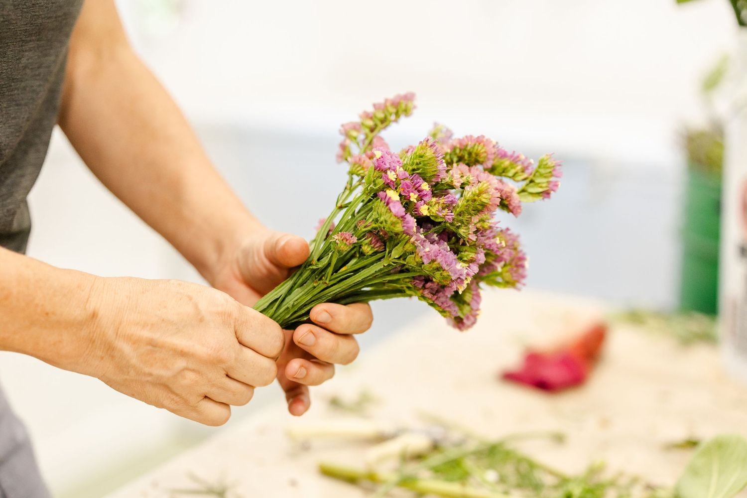 Hands arranging freshly cut flowers and produce on a light colored surface.