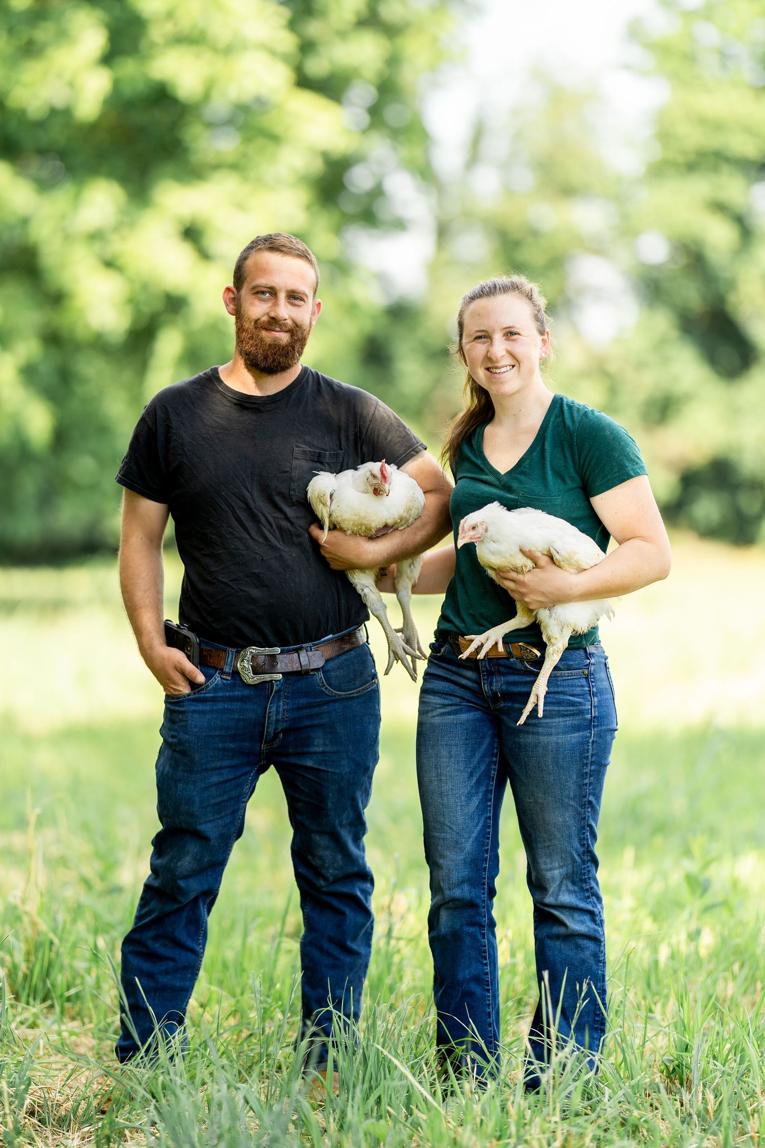 Two farmers holding white chickens while standing in sunny green field.