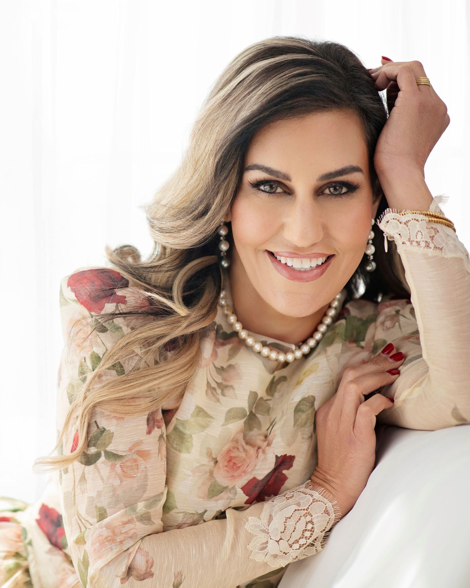 Smiling woman with blonde hair wearing a floral dress and pearl necklace posing elegantly against a white background.