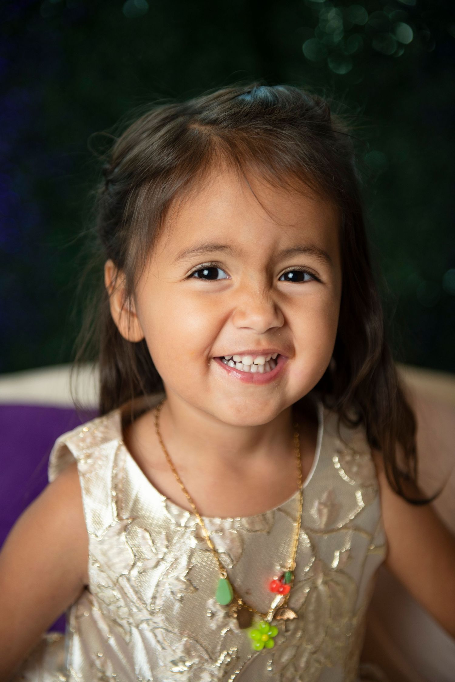 A smiling young girl in a white dress wearing a colorful necklace, posing happily against a dark background.