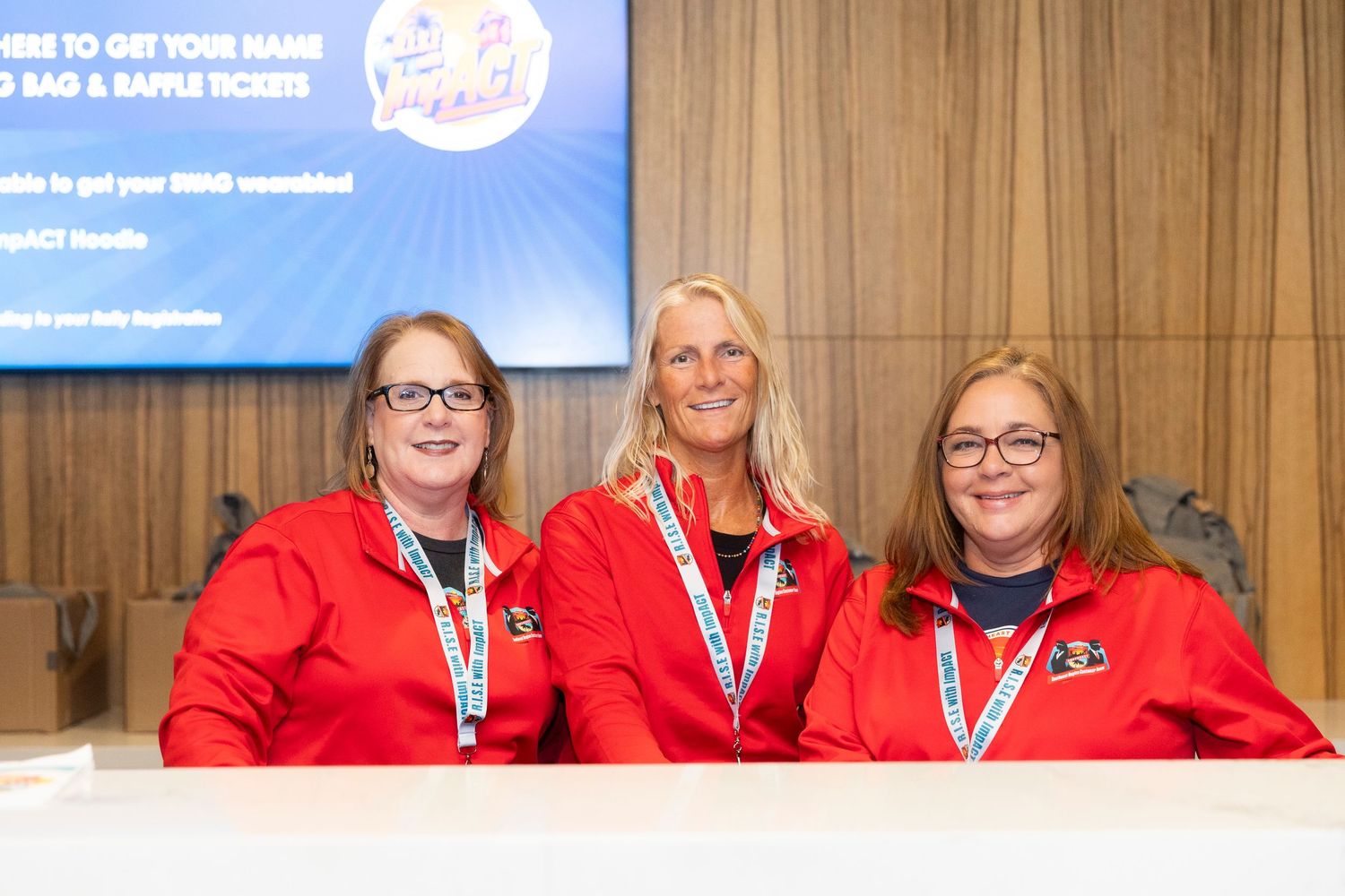 Three women in red jackets smile at a registration table during a corporate event or conference.