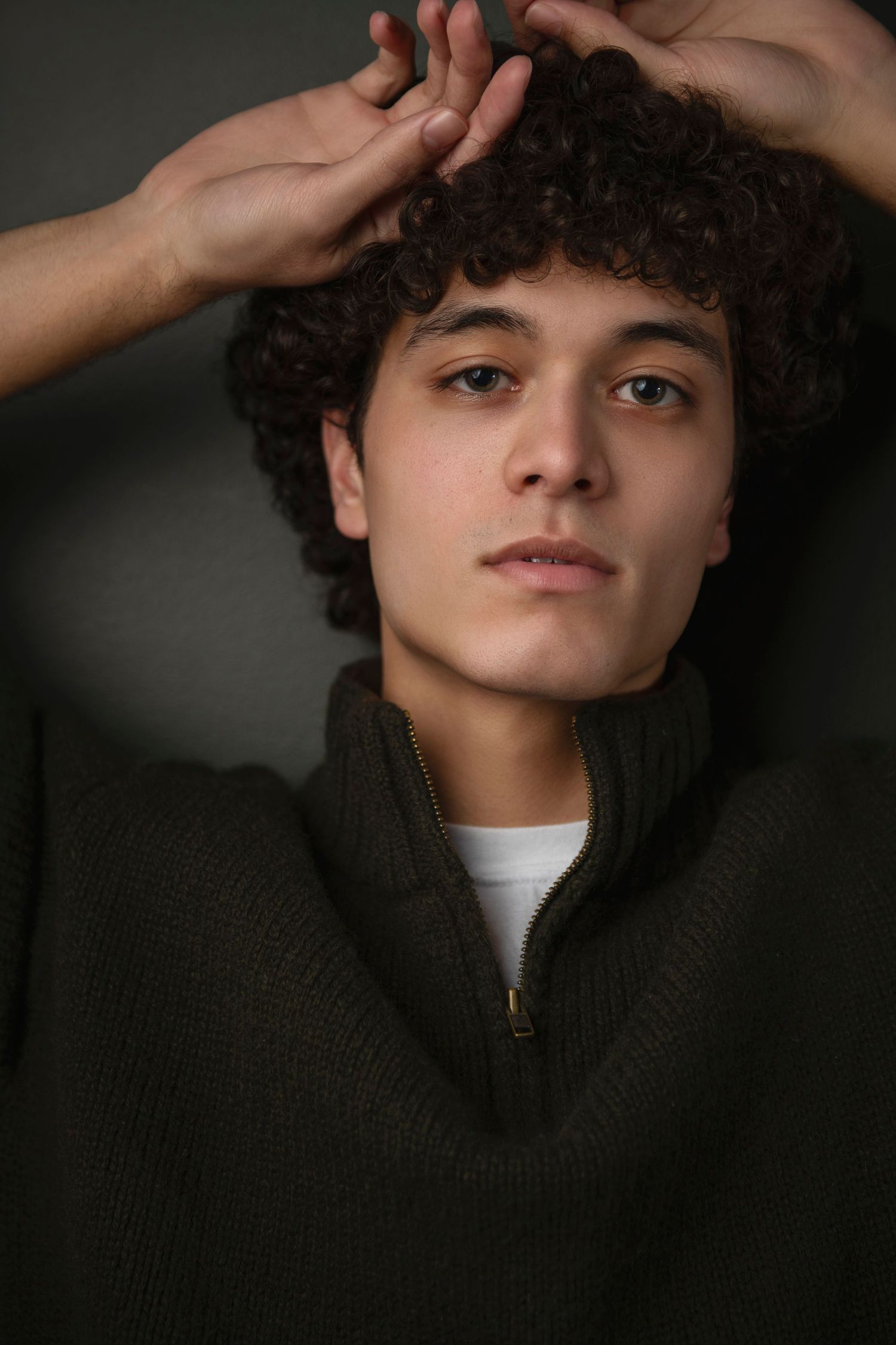 Portrait of a young man with curly dark hair in a green zip-up sweater, hands raised behind his head.