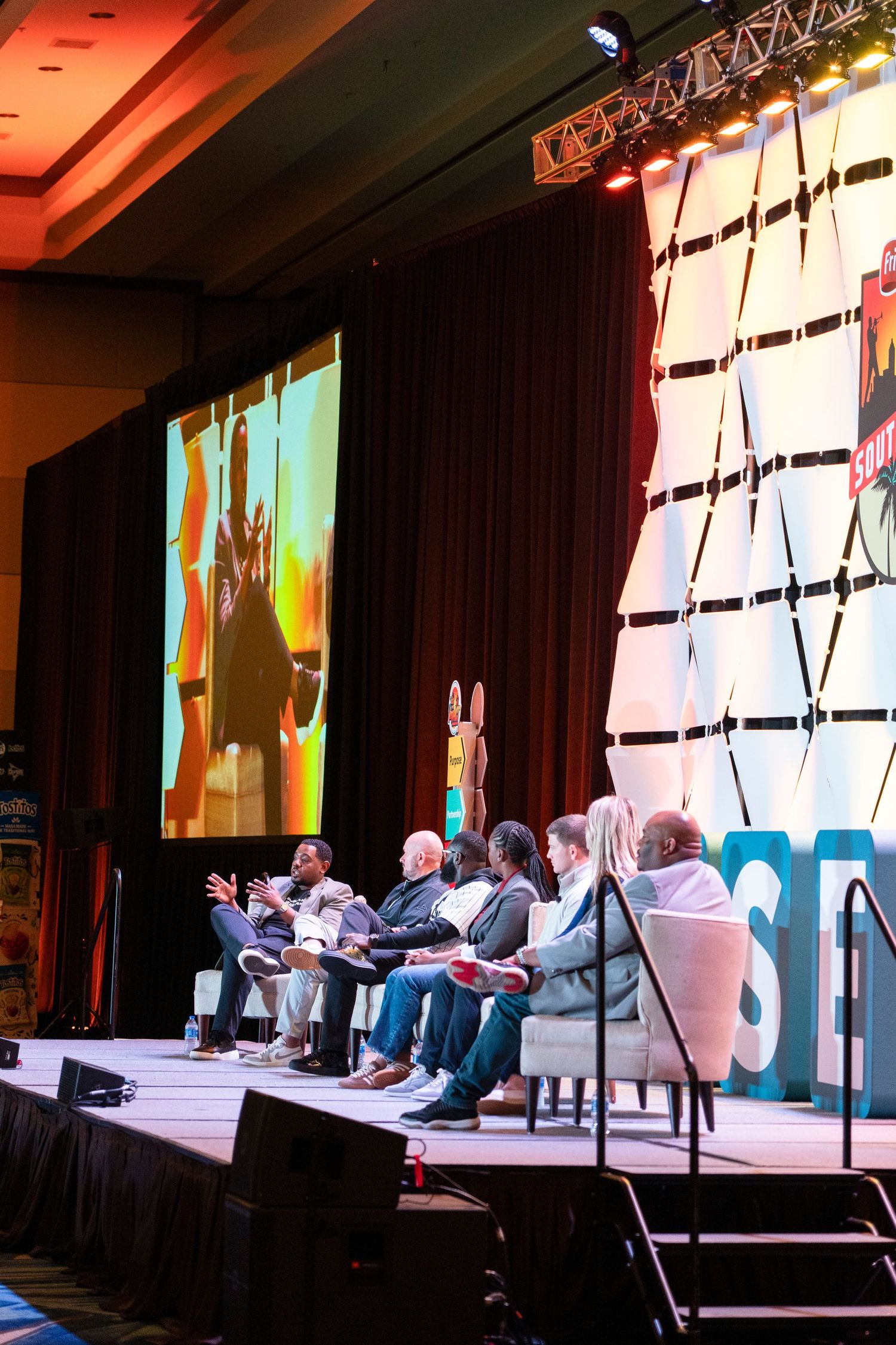 Panel discussion on stage at a conference with speakers seated in chairs, large projection screen and decorative backdrop behind them.