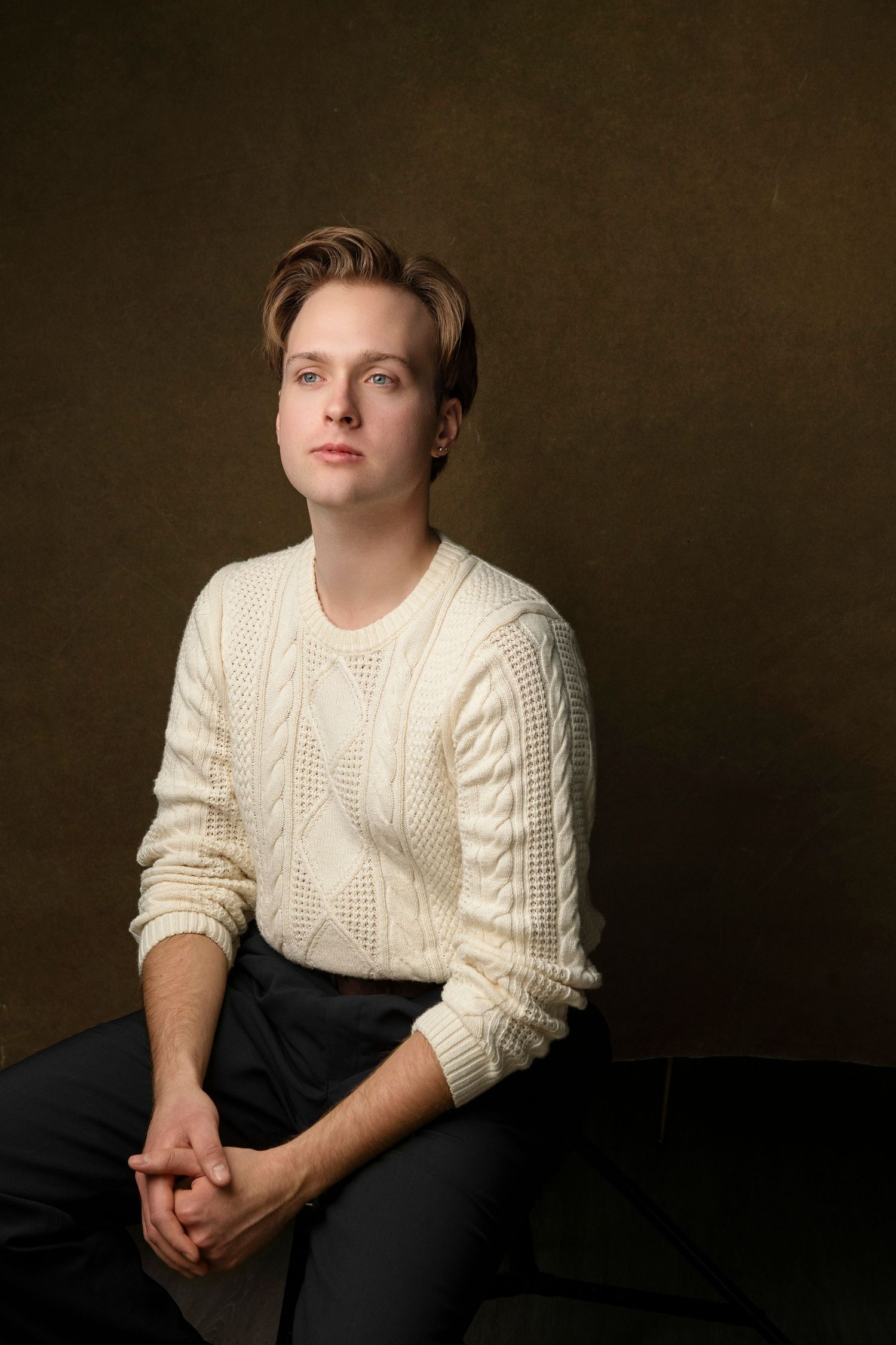 Young man in cream cable-knit sweater and dark pants sitting with clasped hands on dark background.