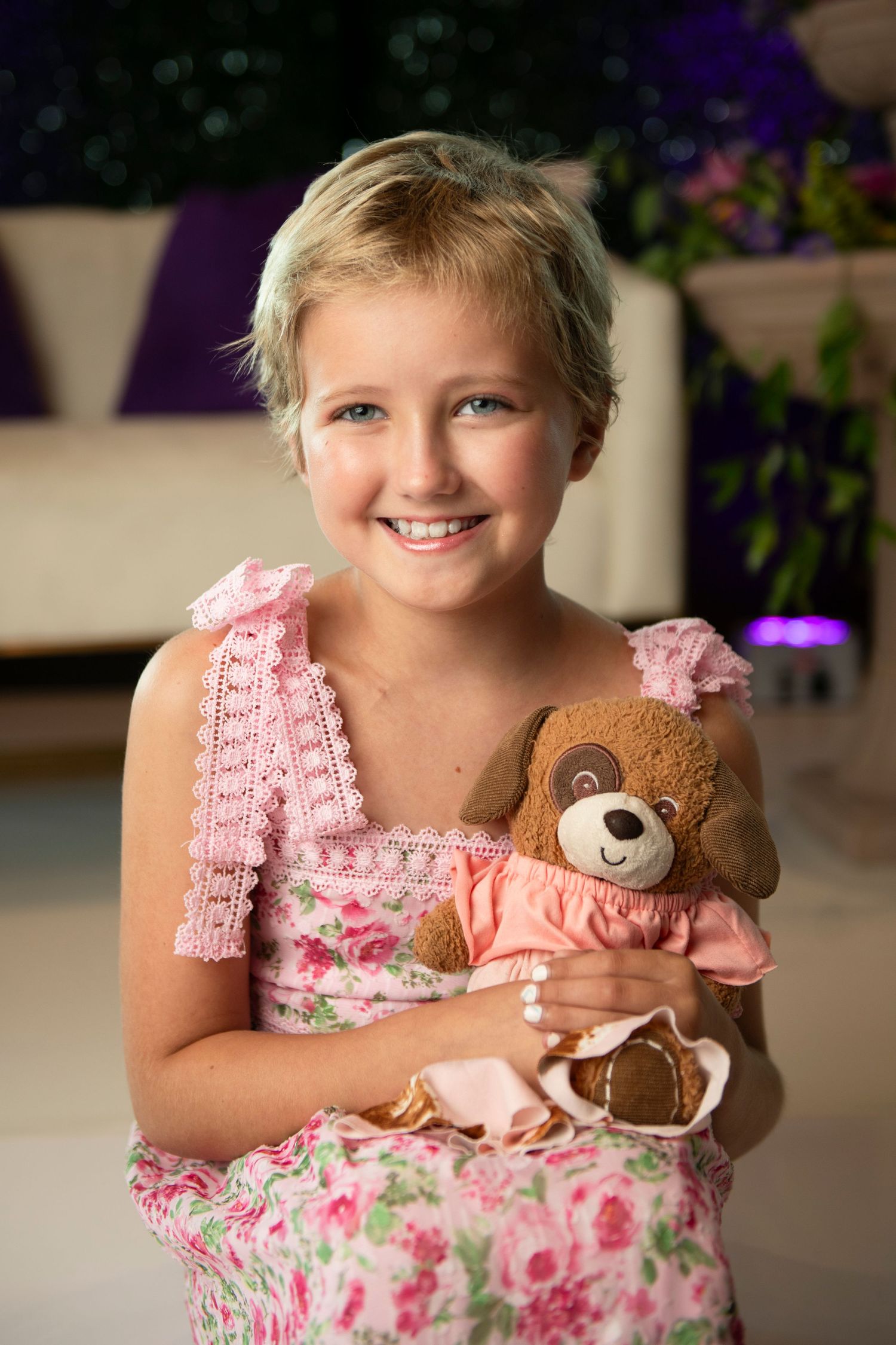 Smiling young girl in pink floral dress holding a brown teddy bear in a pink outfit.