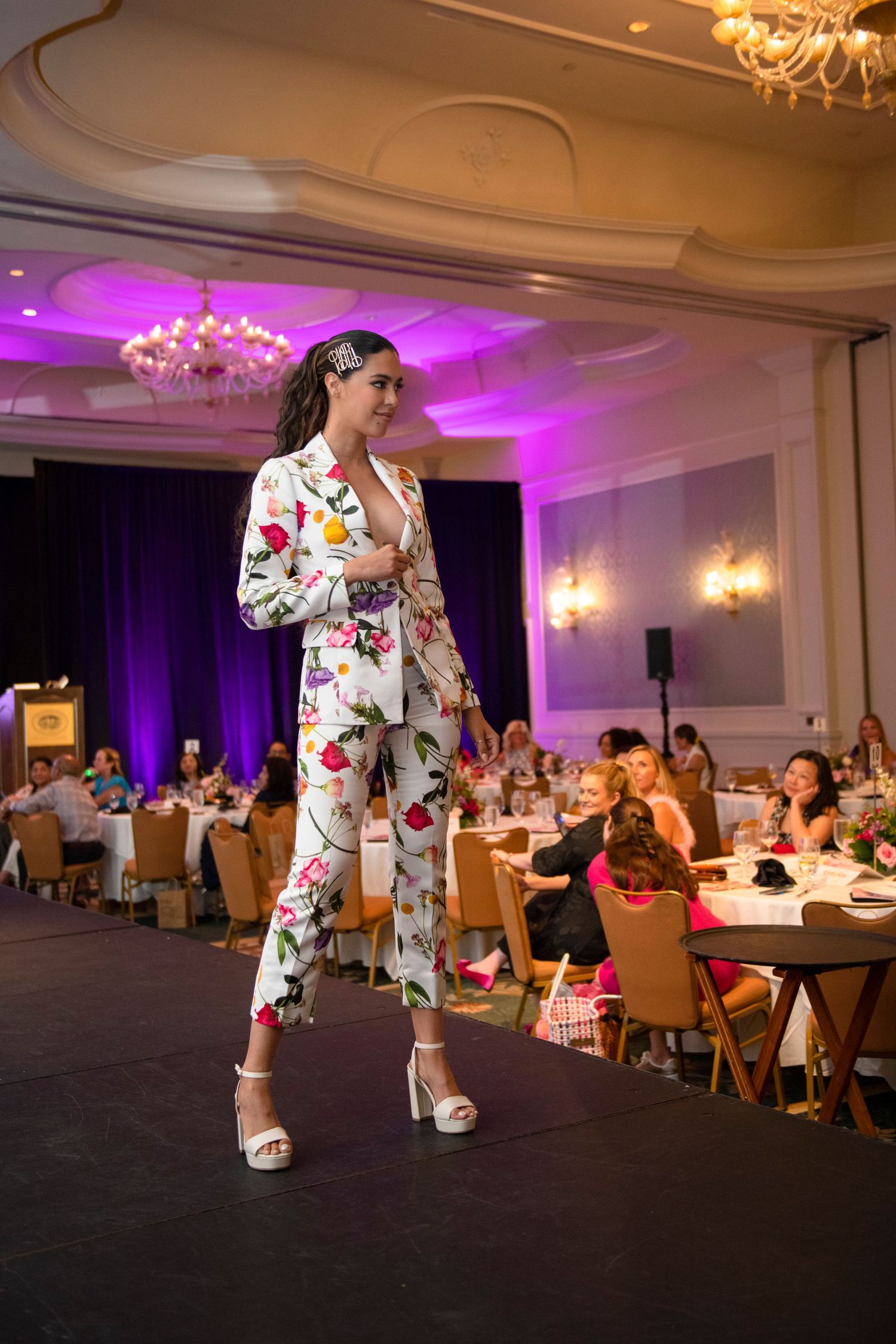 Model walks runway in floral white pantsuit at elegant ballroom fashion show with purple lighting.