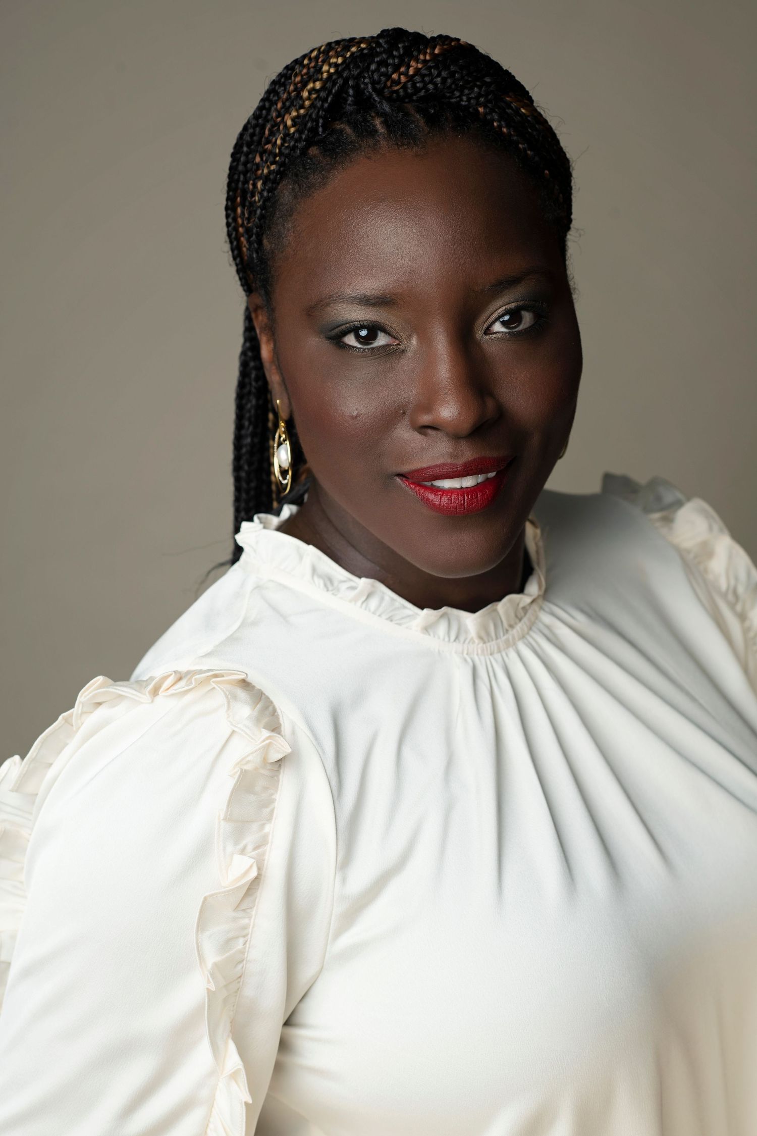 Confident Black woman with braids, red lips, gold earrings, wearing elegant white ruffled blouse, studio portrait.