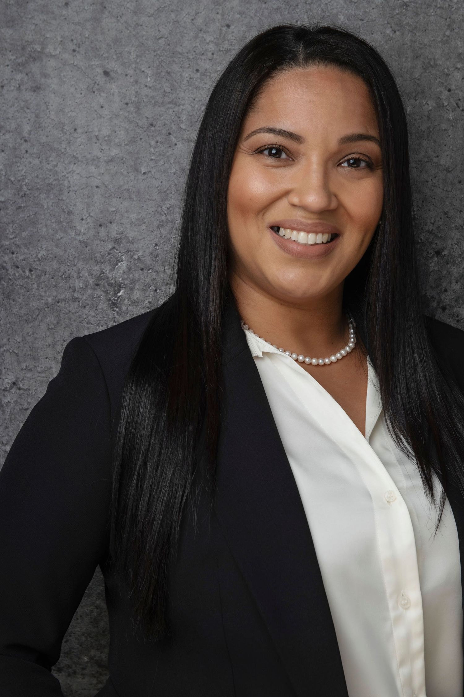 Professional headshot of a smiling woman in a black blazer and pearl necklace against a gray background.