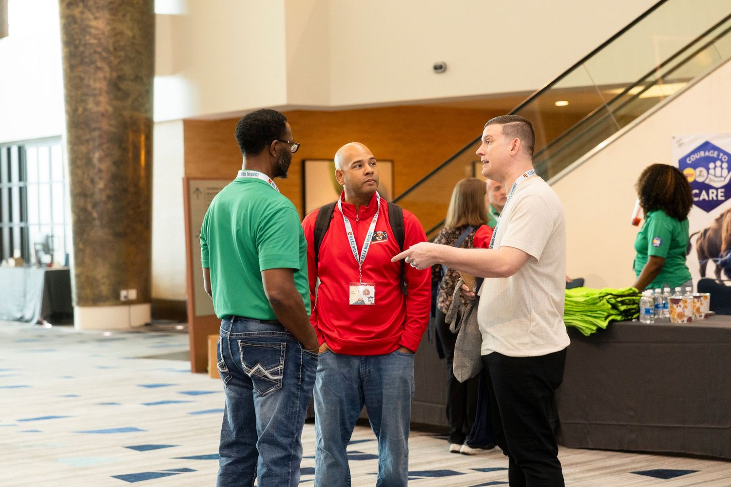 Three professionals networking at a conference event in a modern venue lobby with exhibitor booths in background.