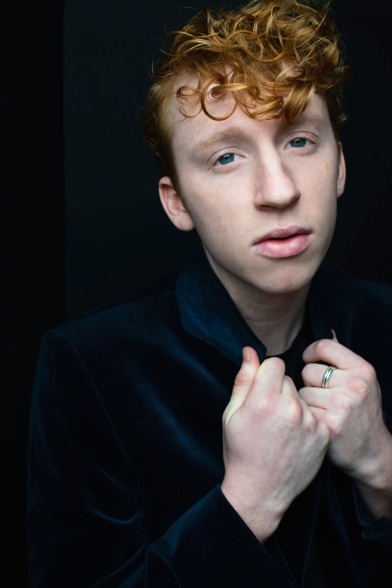 Young man with red curly hair in dark velvet jacket holds collar while gazing at camera.