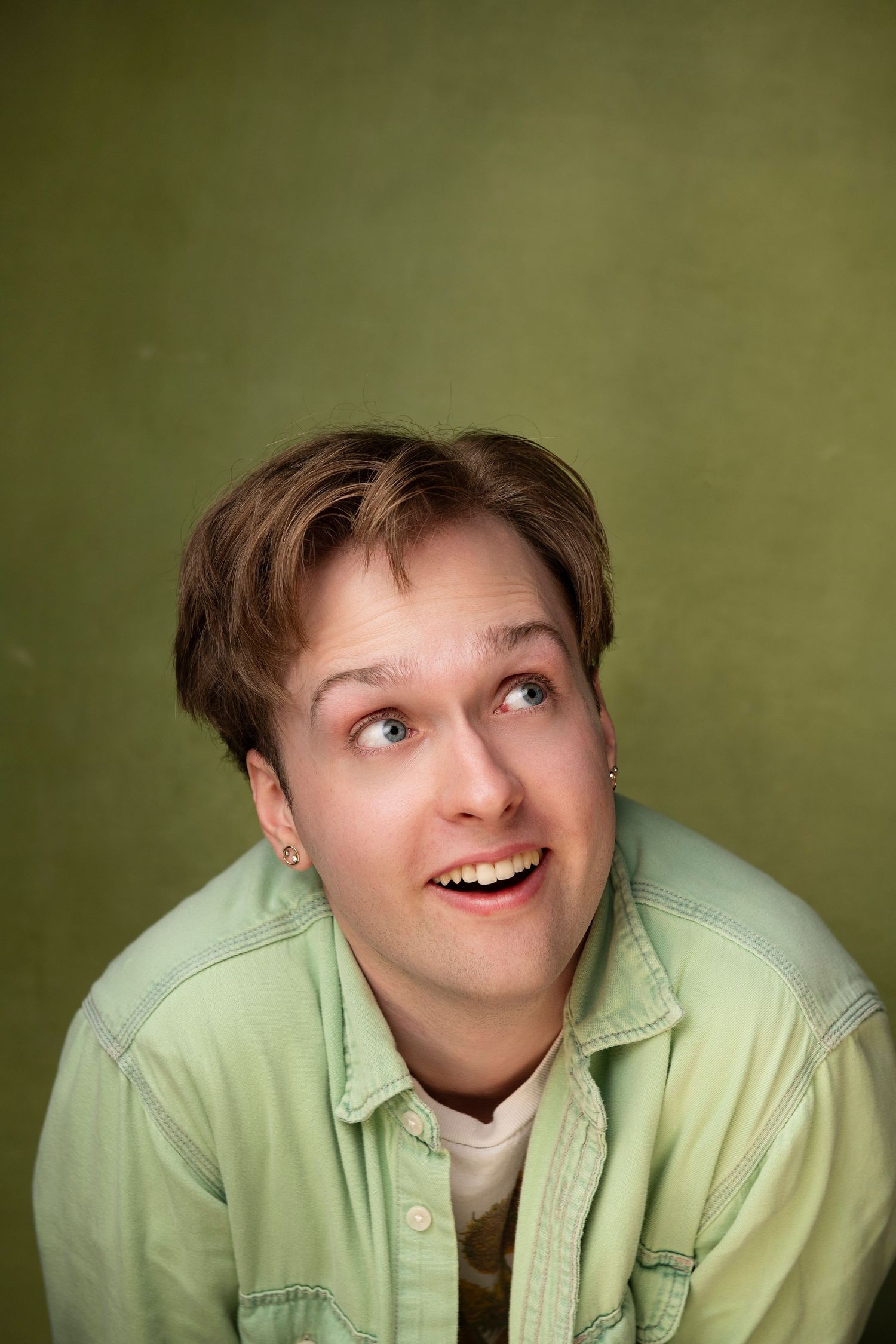 Young man in light green denim jacket looking up and smiling against an olive green background.