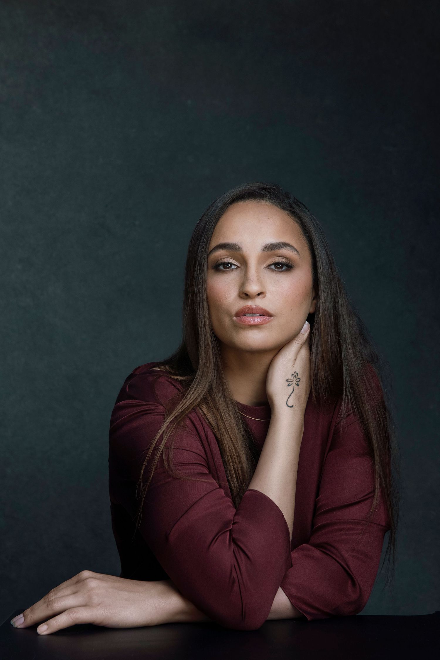 Young woman in burgundy top with hand tattoo poses thoughtfully at table against dark teal background.