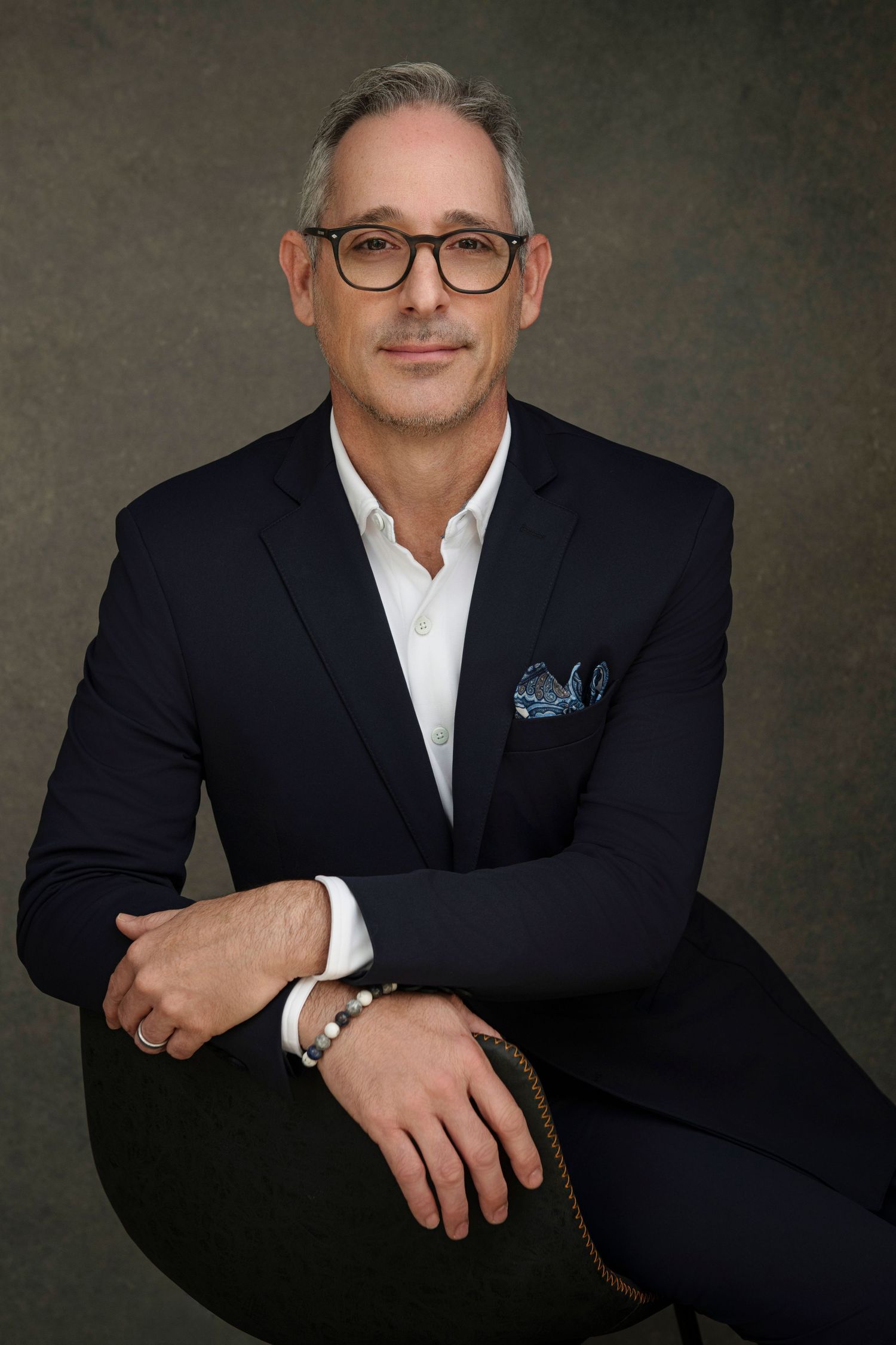 Professional man in navy suit with pocket square and glasses poses confidently for a business portrait photo.