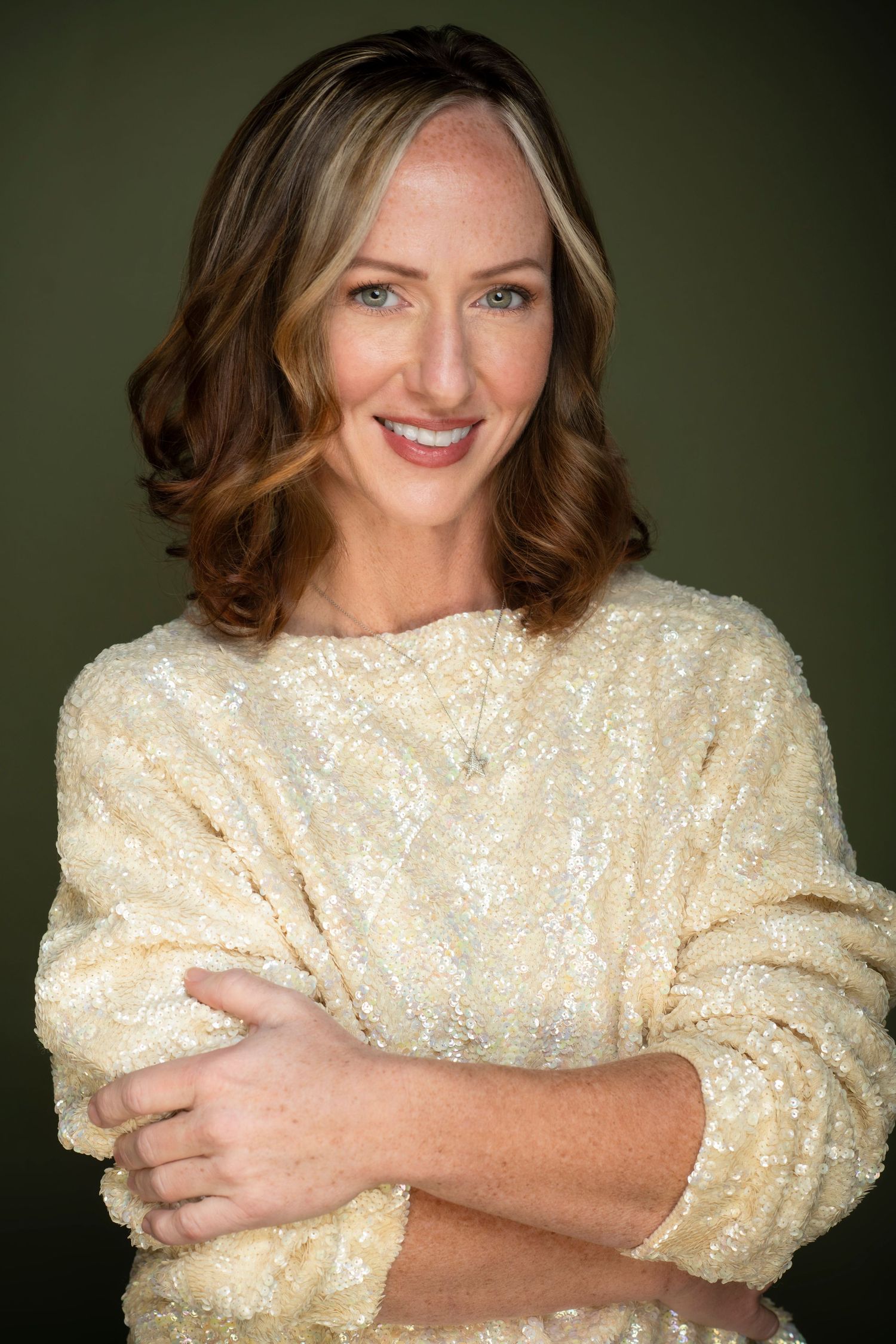 Smiling woman with auburn hair wearing a cream sequined sweater, arms crossed, posing against a dark green background.