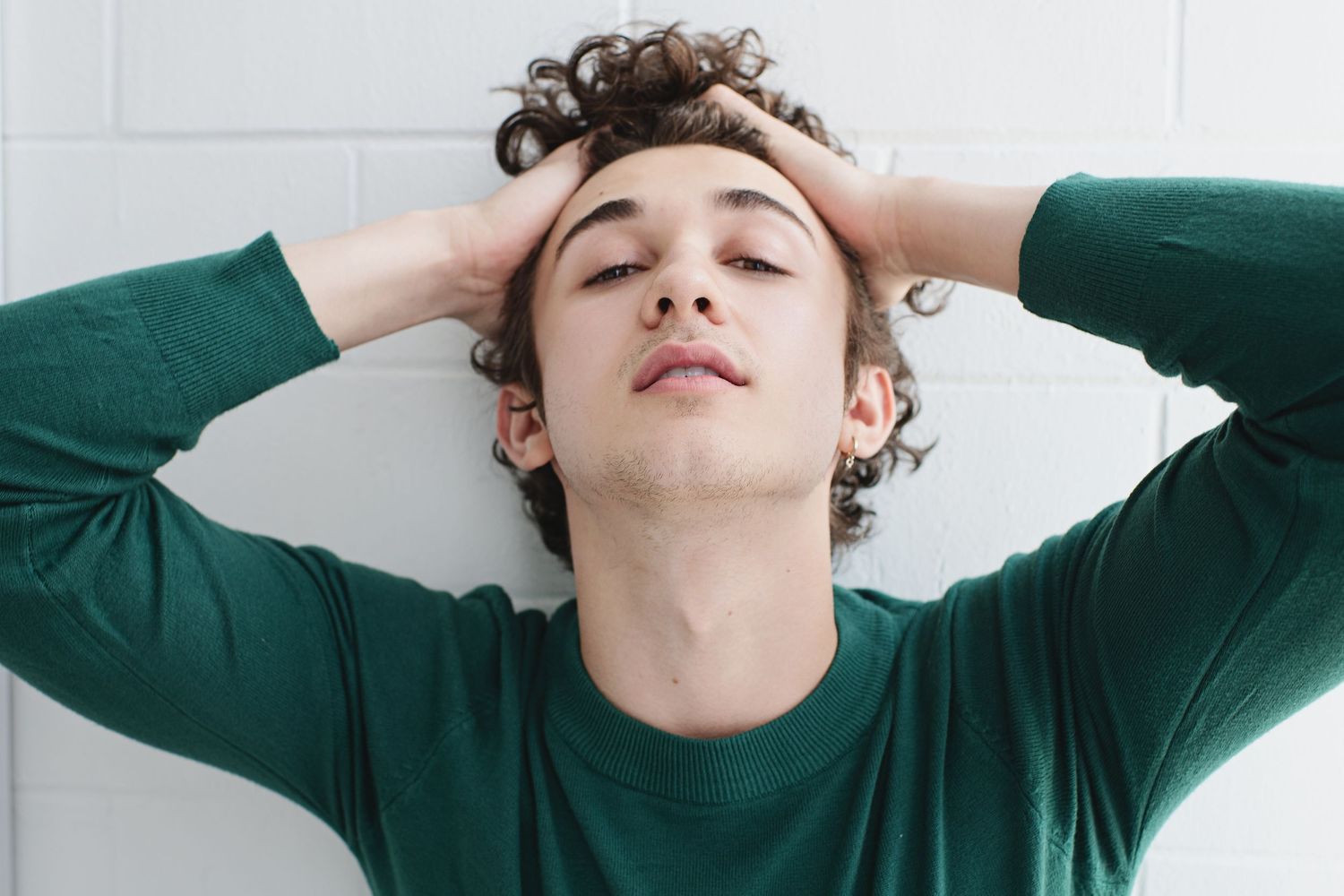 Young man in green long-sleeve shirt with hands on head, eyes closed, leaning against white wall.