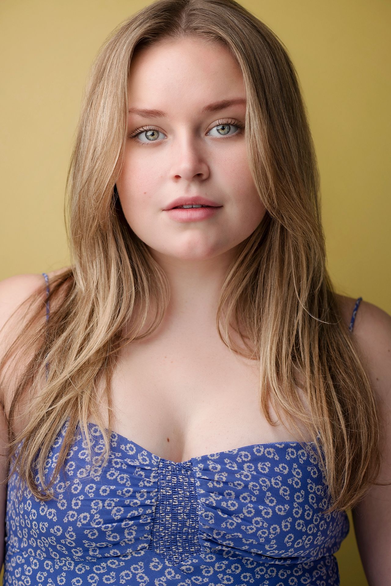 Young blonde woman with green eyes wearing a blue floral top poses against a yellow background.