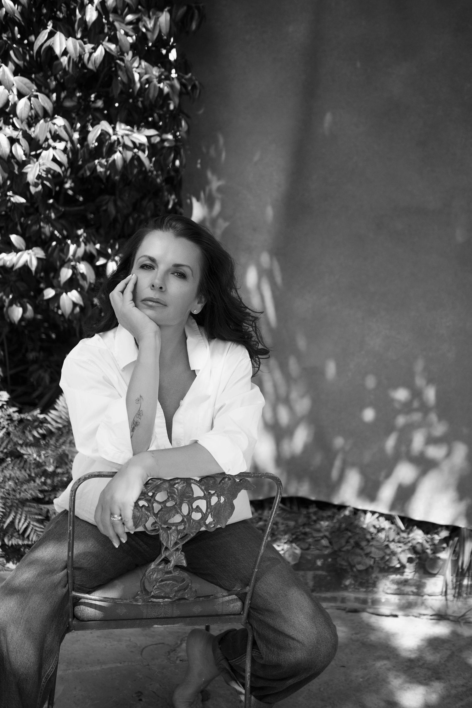 Black and white photo of a woman in white shirt and jeans sitting on a chair outdoors near lush green foliage.