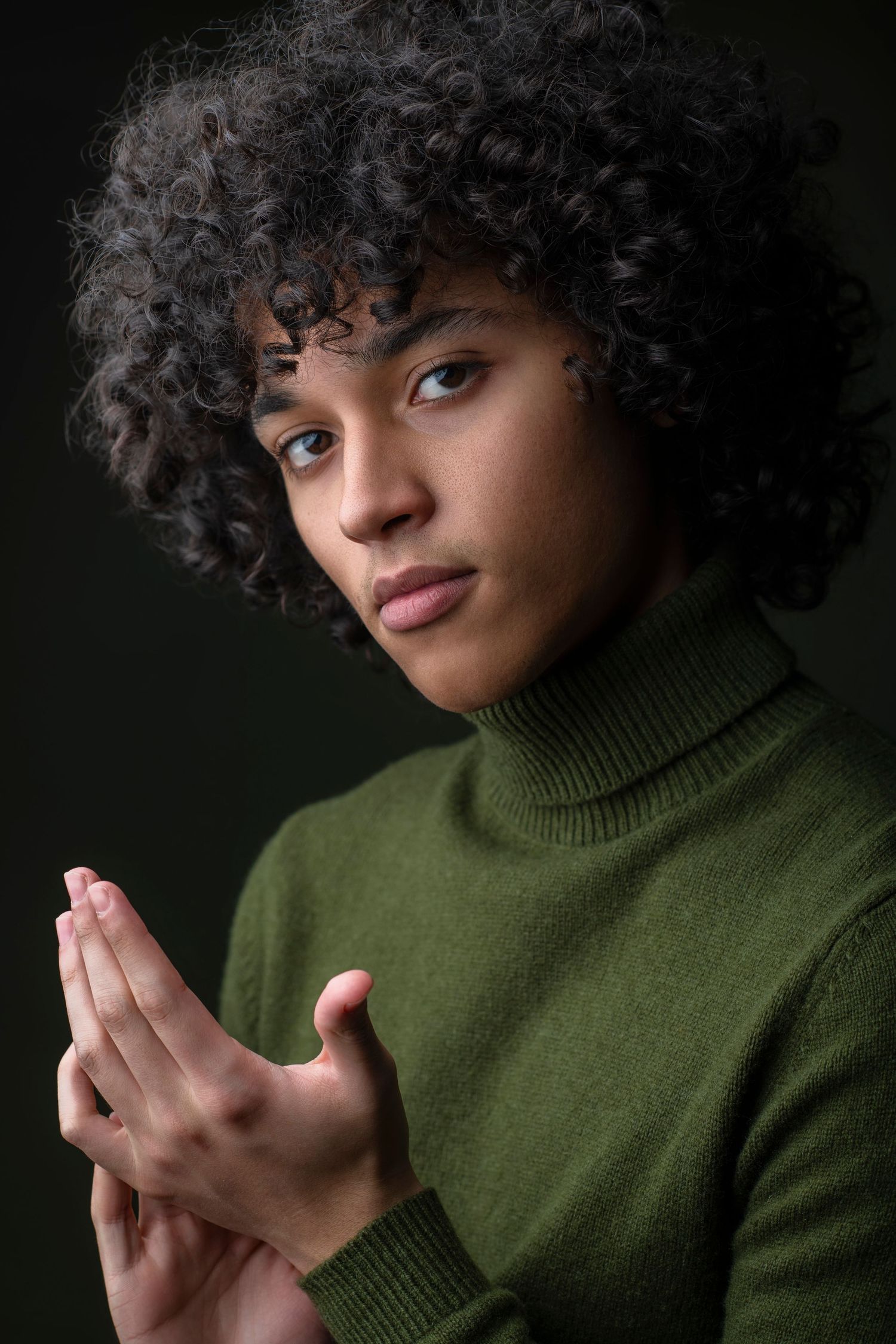 Person with curly afro hair in green turtleneck sweater pressing hands together against dark background.