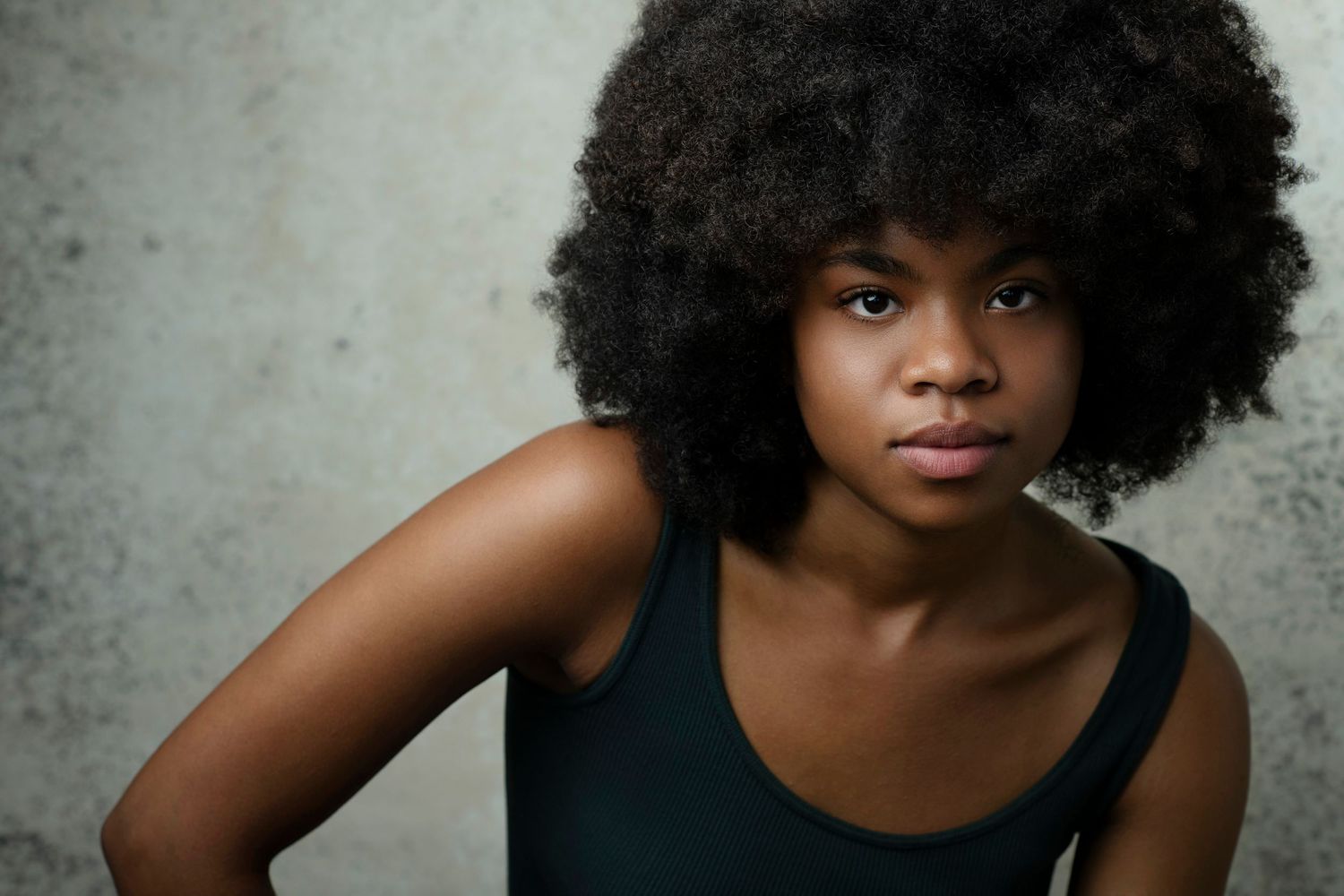 Young Black woman with natural afro hairstyle wearing a dark tank top, posing confidently against a gray background.
