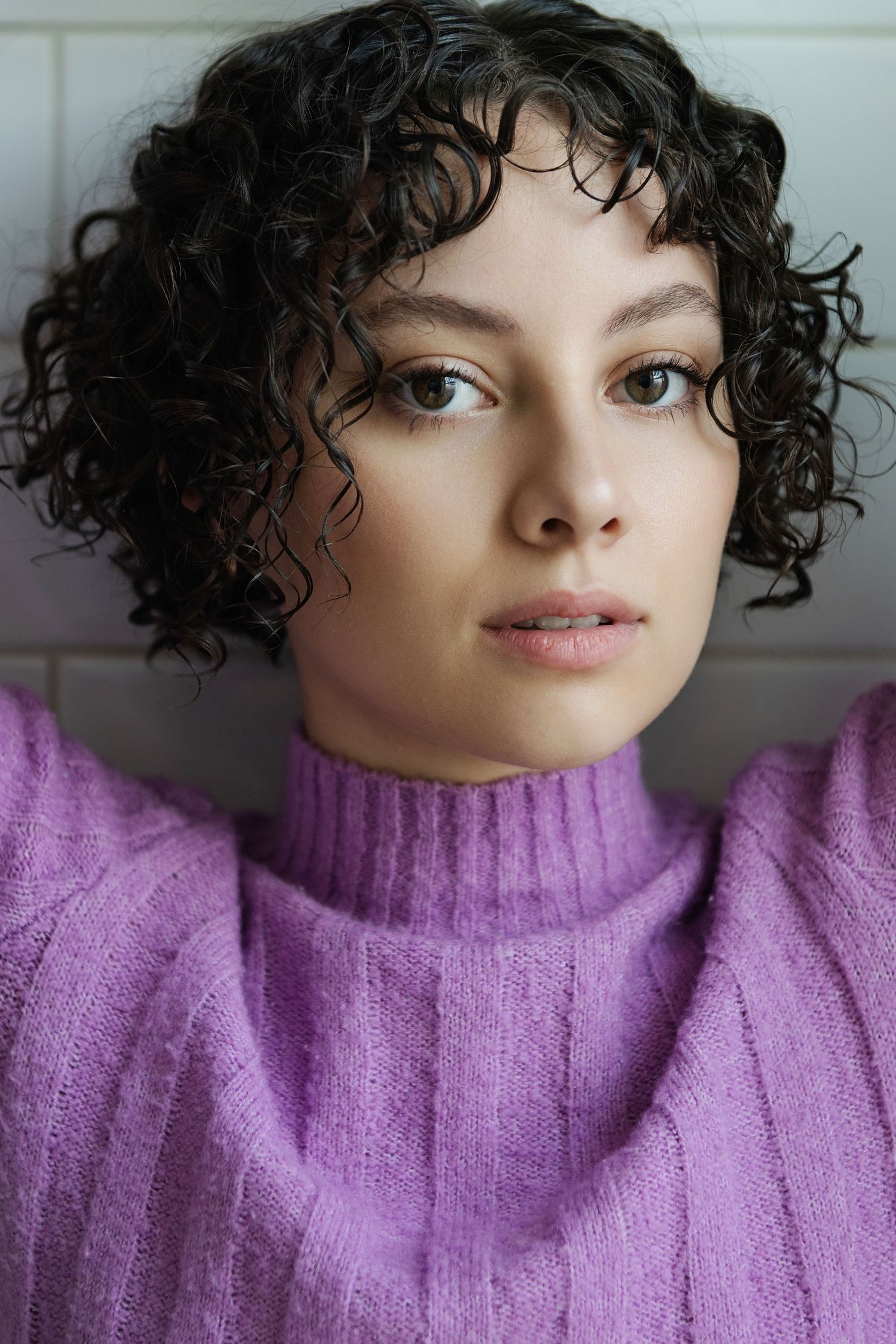 Young woman with short curly dark hair wearing a purple turtleneck sweater, posing against a white tiled wall.