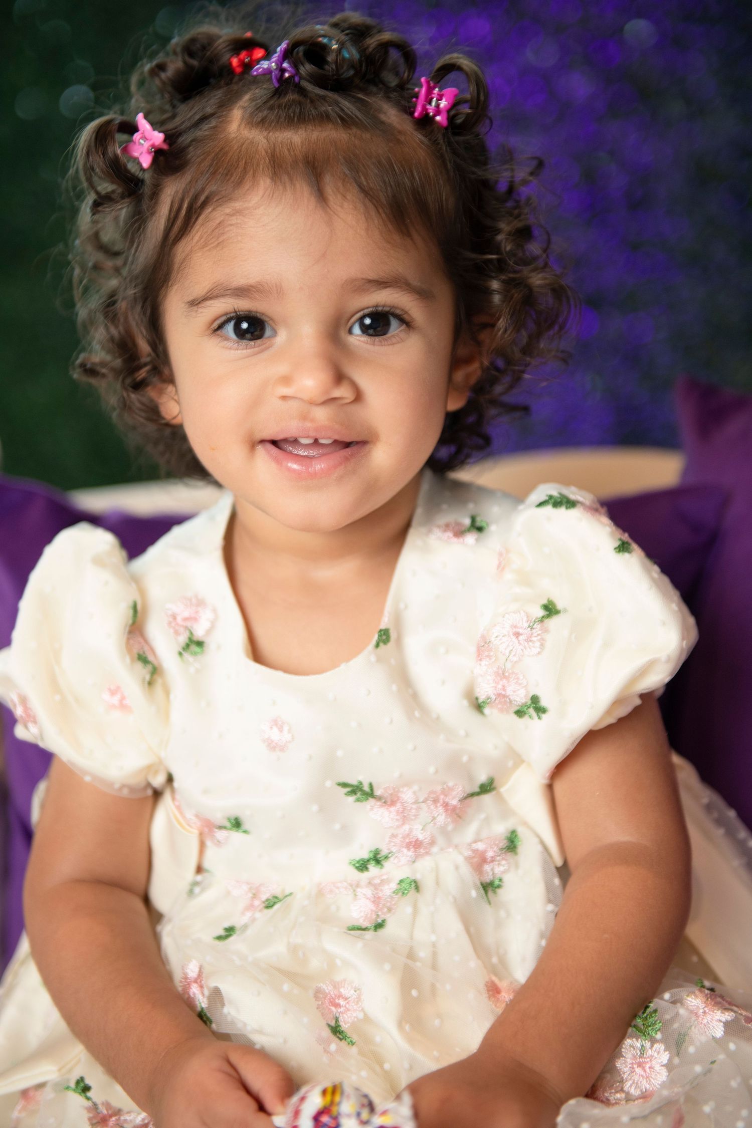 Adorable toddler girl in floral white dress with curly hair and colorful clips, smiling at camera.