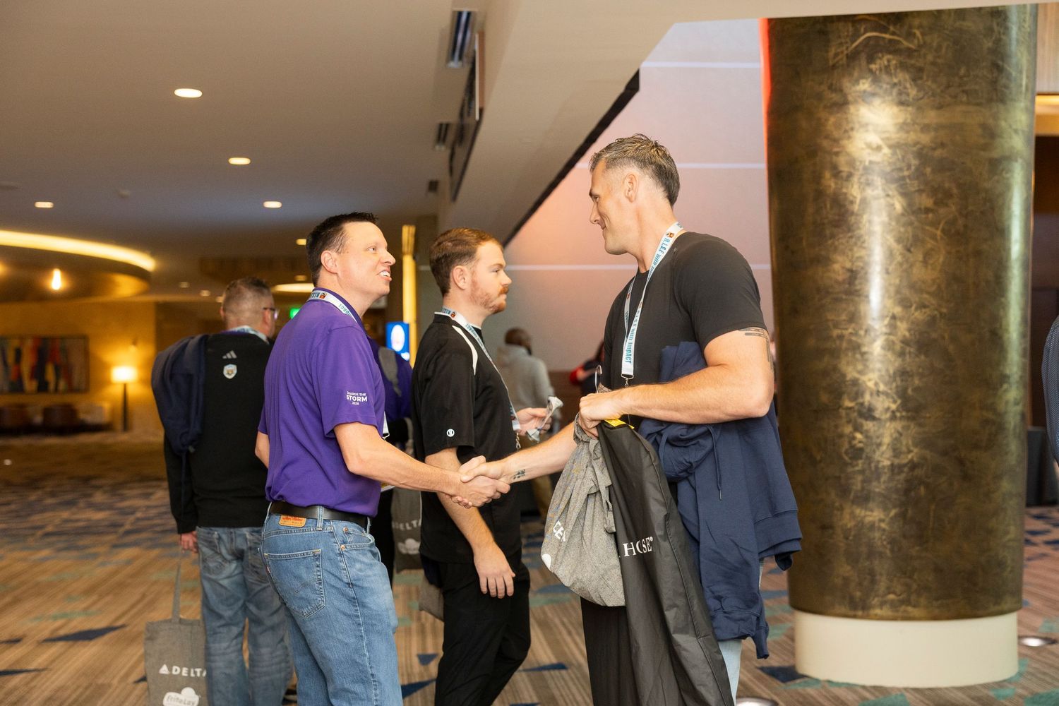Three professionals networking and shaking hands in a modern conference hall lobby near decorative columns.
