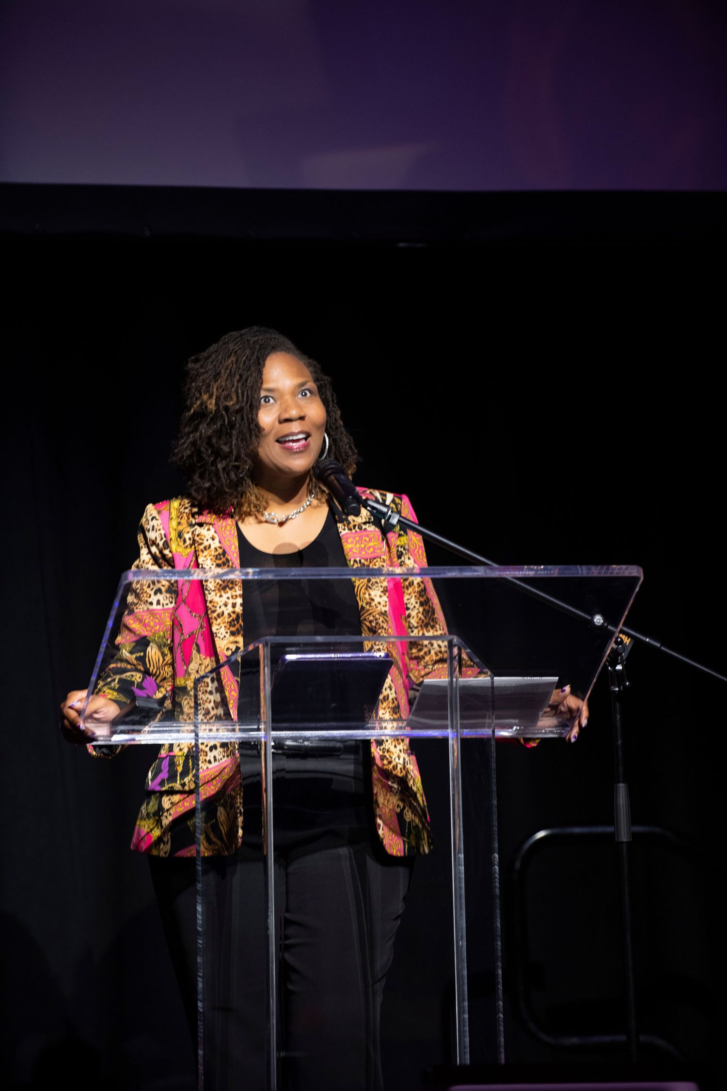 Woman in colorful blazer speaking at a clear podium on a dark stage during a formal event.