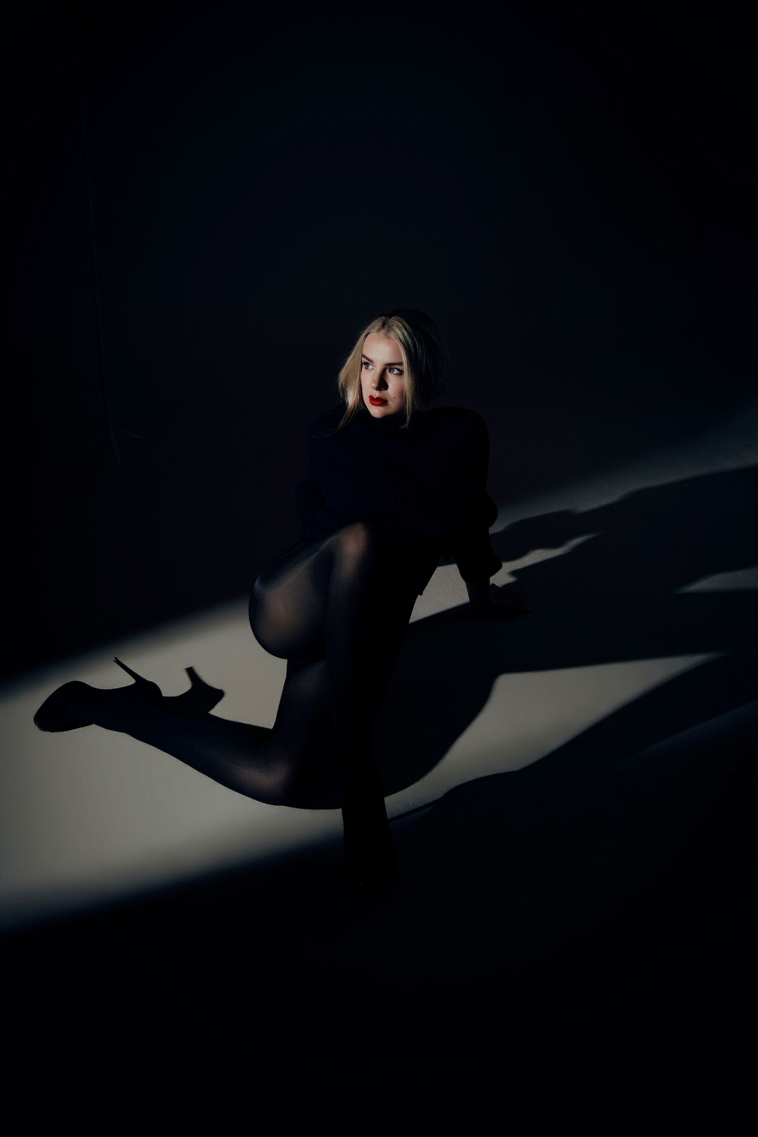 Blonde woman with red lipstick poses on the floor in dark tights and high heels, lit by dramatic beams of light and sharp shadows.