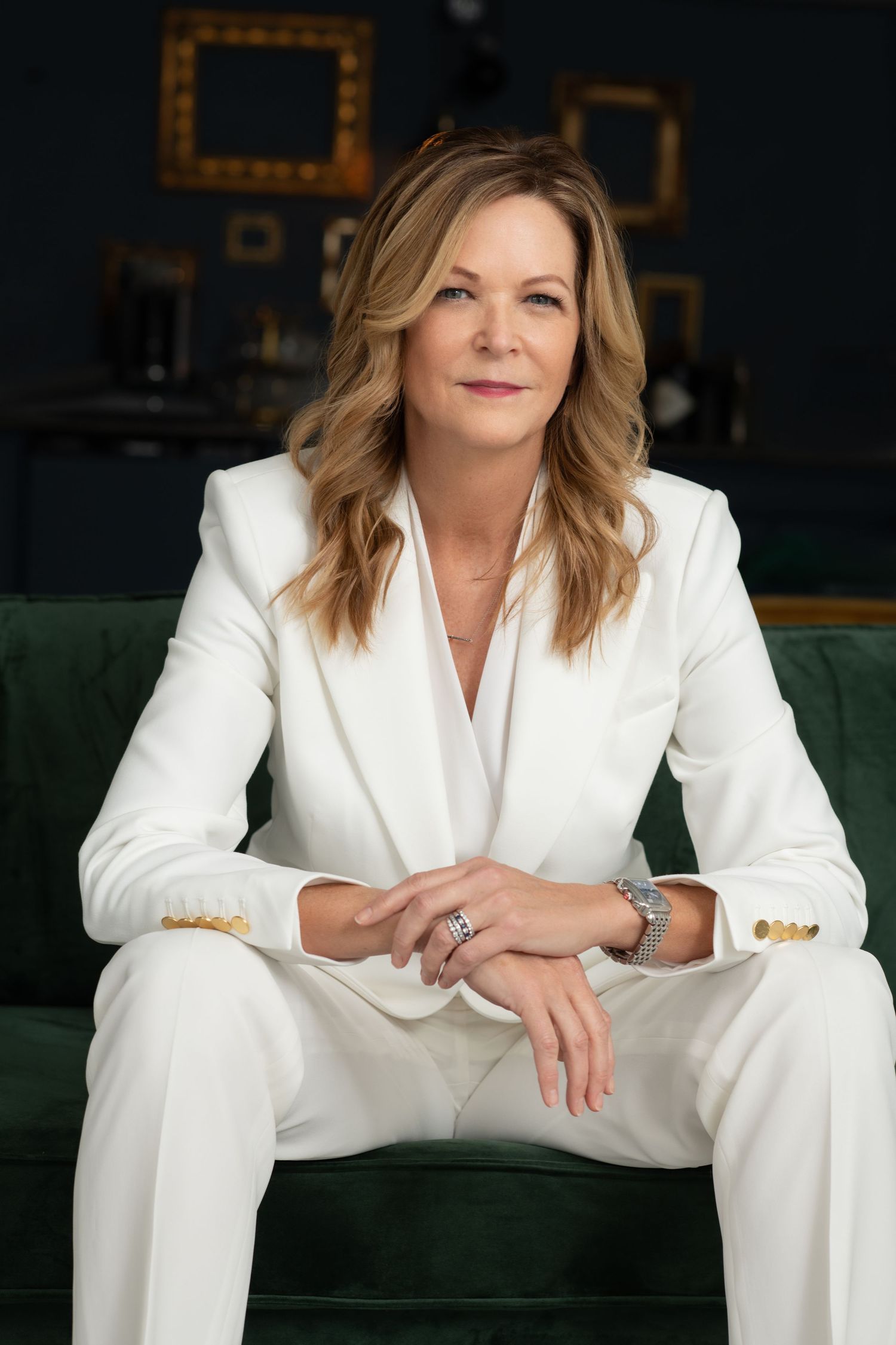 A confident woman in a white suit sits on a green velvet sofa, with gold-framed art in the background.
