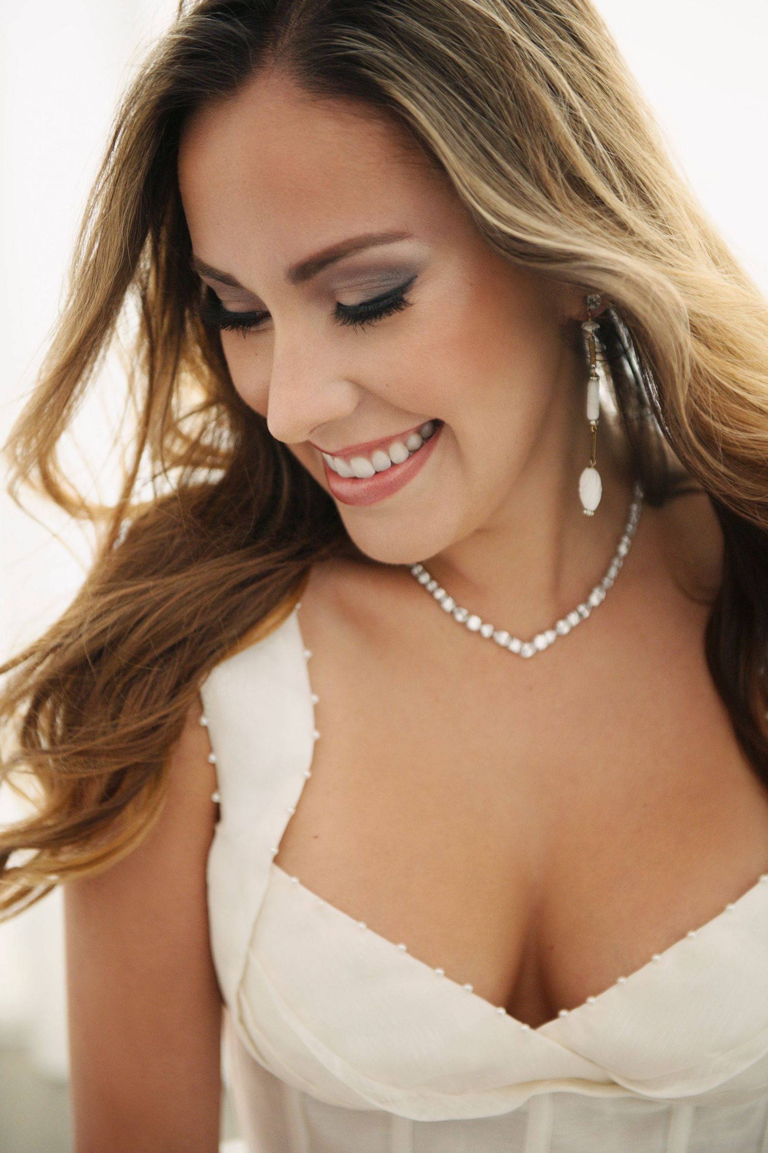 Smiling brunette woman wearing pearl necklace and earrings with white dress, looking down elegantly.
