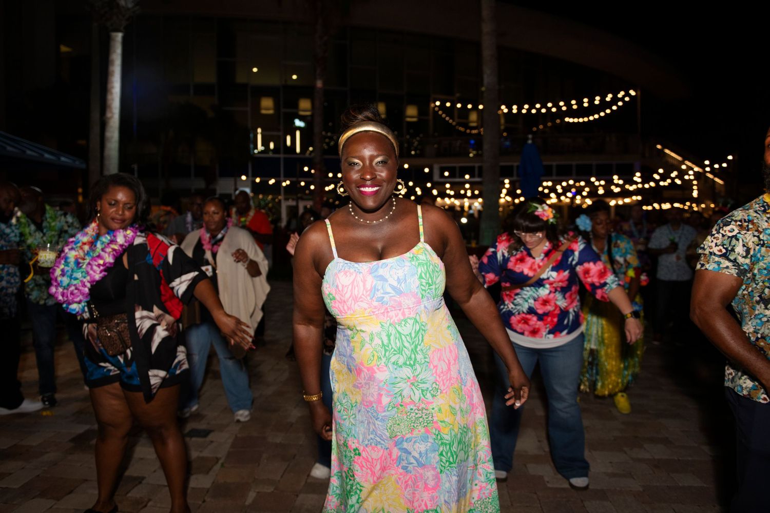 Woman in colorful floral dress smiling and dancing outdoors at a festive night event with string lights.