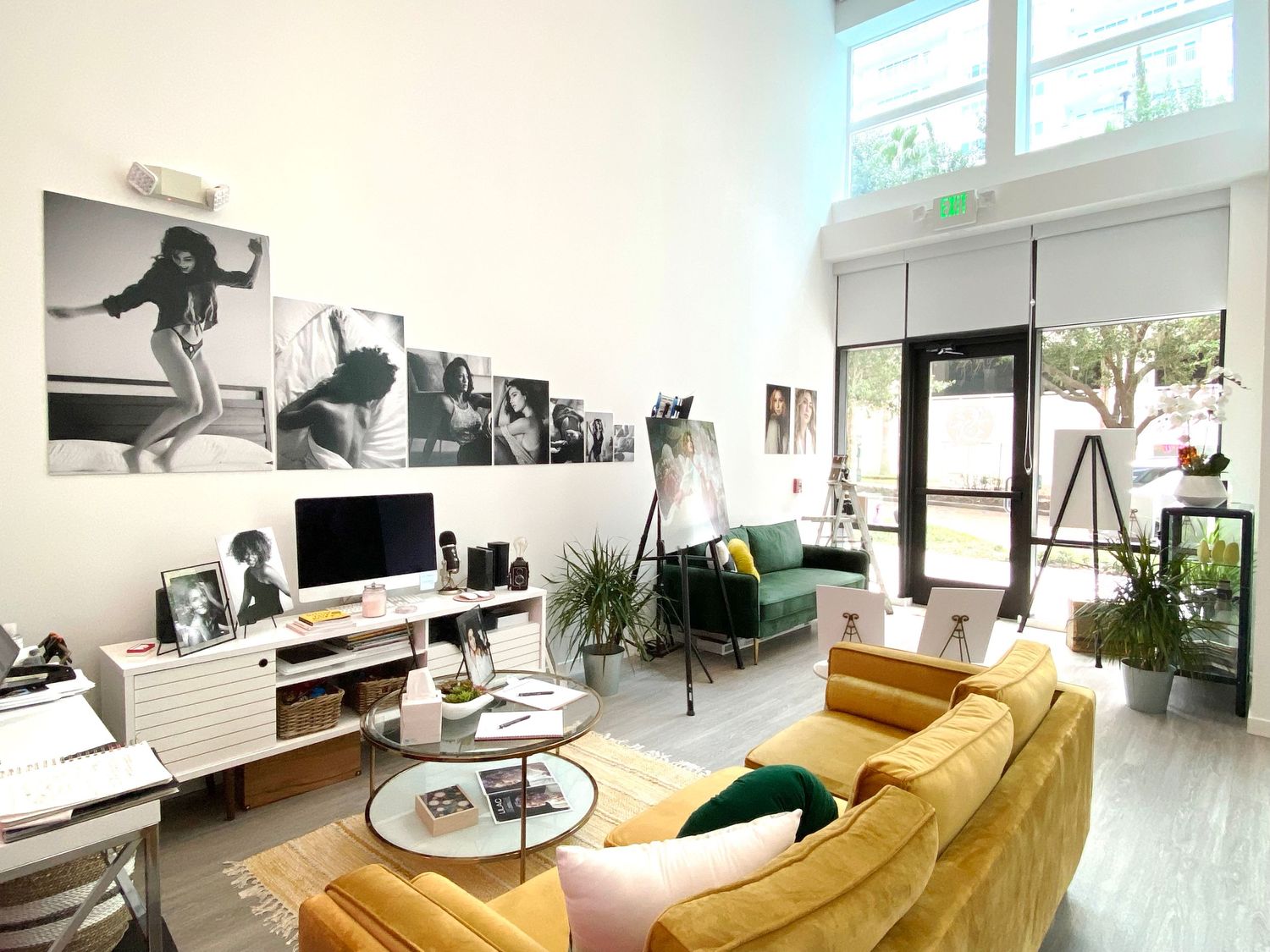 Bright loft living room with a mustard velvet sectional, round glass coffee table, black-and-white art prints, desk setup and green plants.