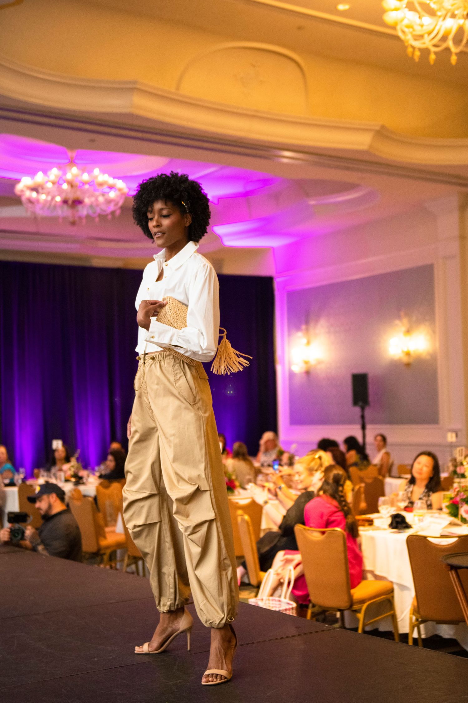 A model walks the runway in gold cargo pants and white blouse at an elegant ballroom fashion show.
