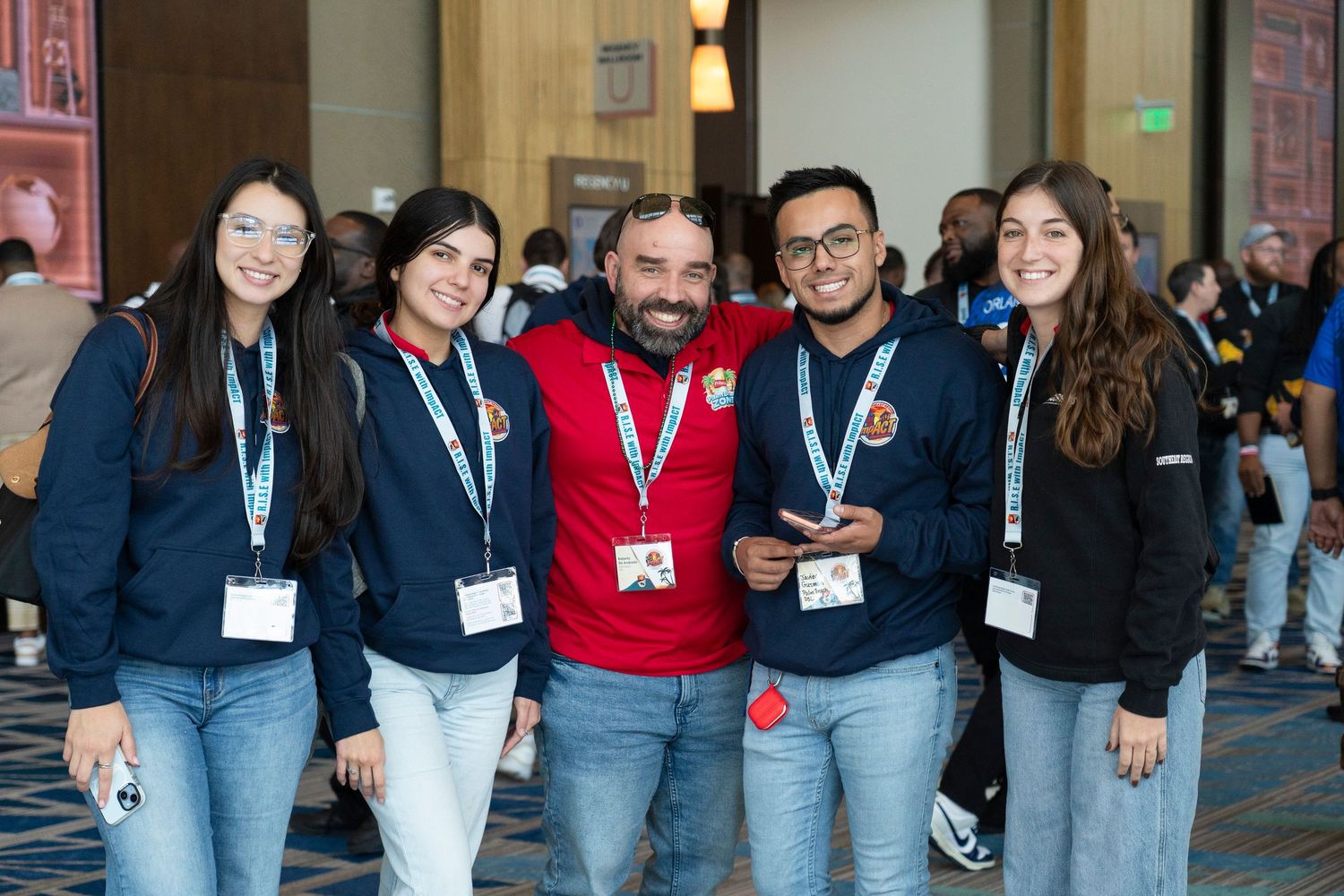 Five smiling conference attendees wearing lanyards and navy jackets pose together in a convention center hallway.