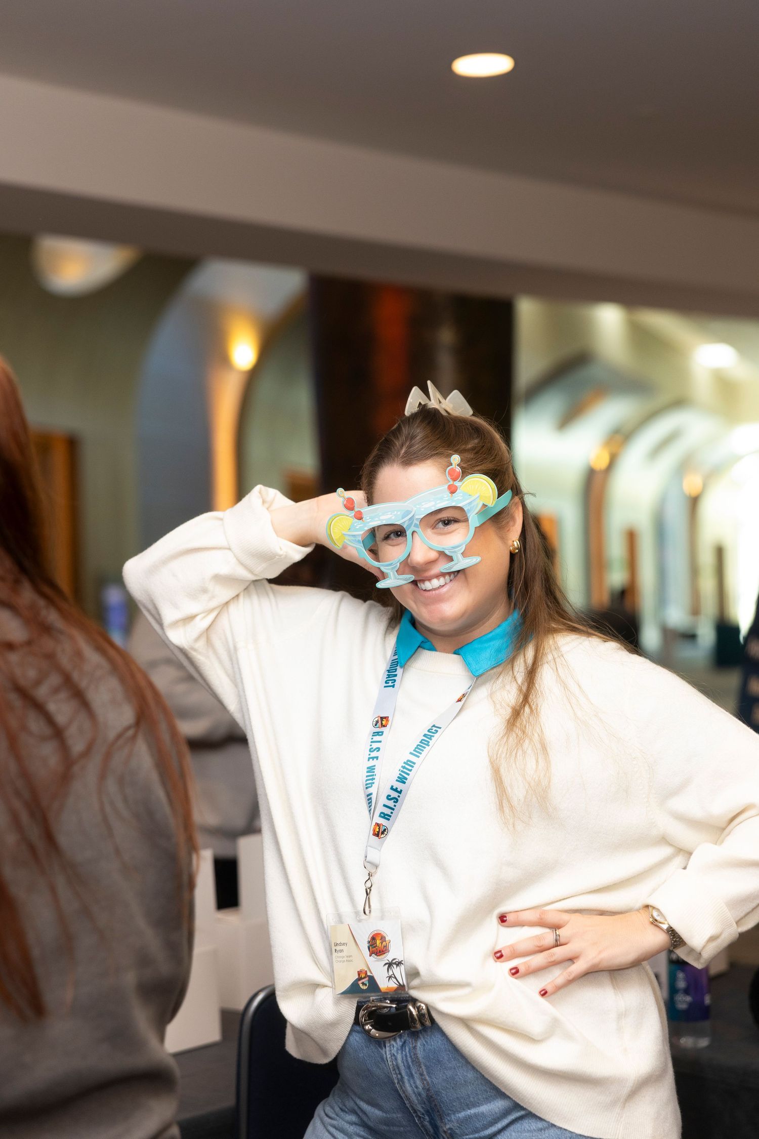 A smiling young woman poses playfully wearing colorful novelty glasses, a white sweatshirt, and a lanyard indoors.