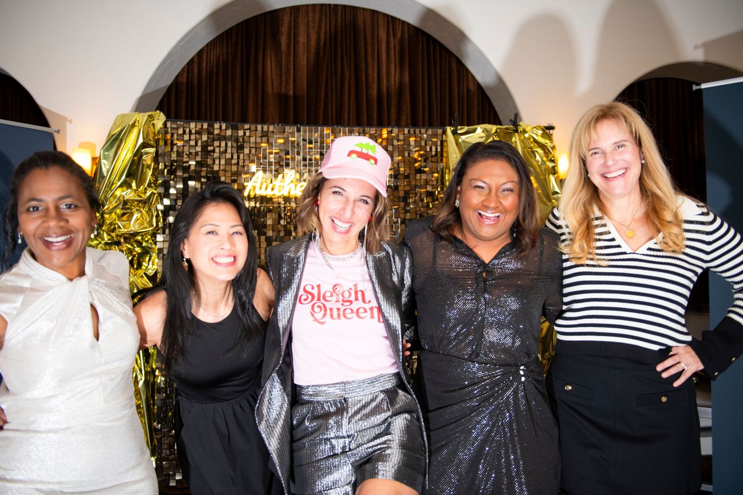 Five women smiling and posing together at a glamorous gold-themed party event.