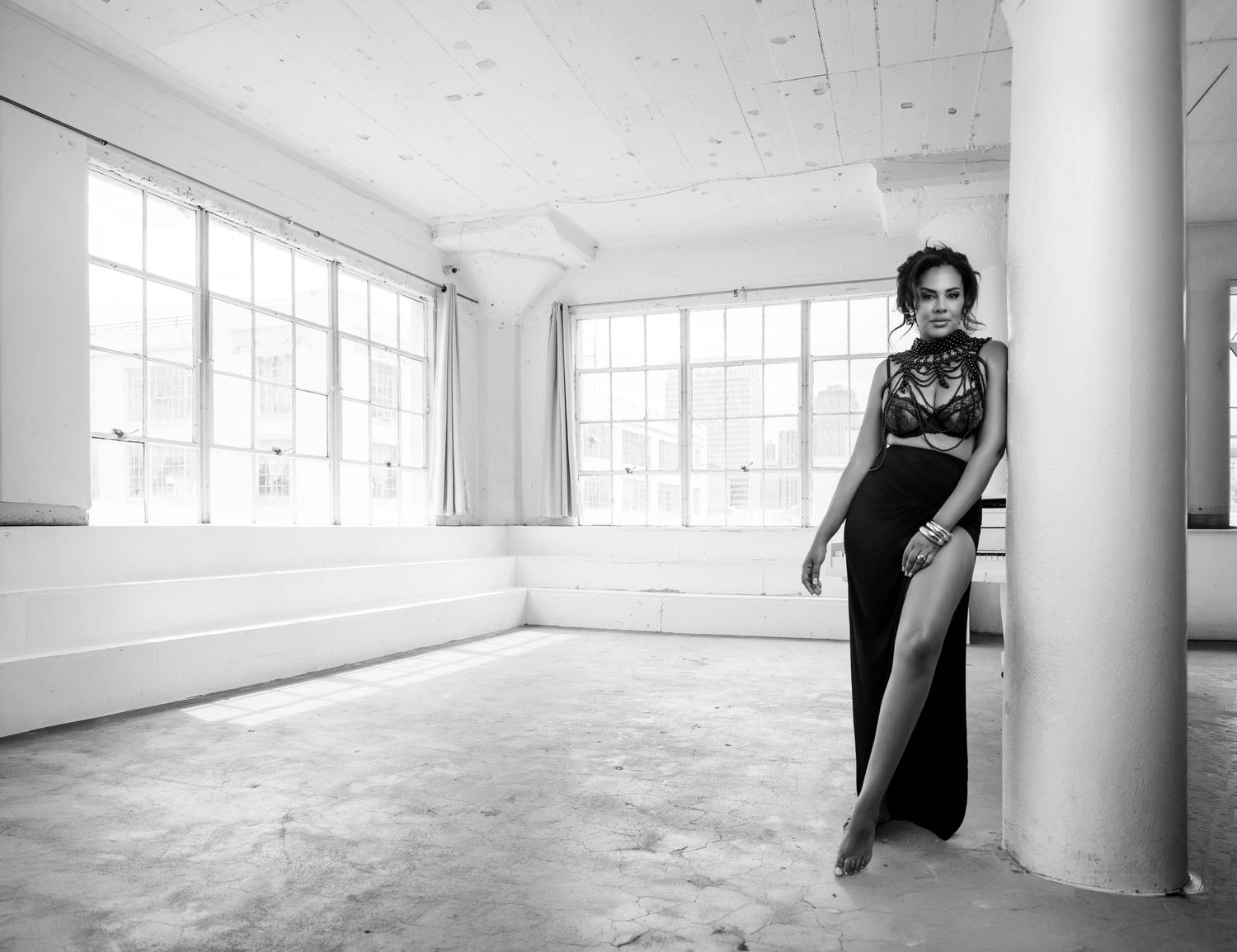 Woman in elegant black gown poses gracefully in bright white studio space with large windows, black and white photo.