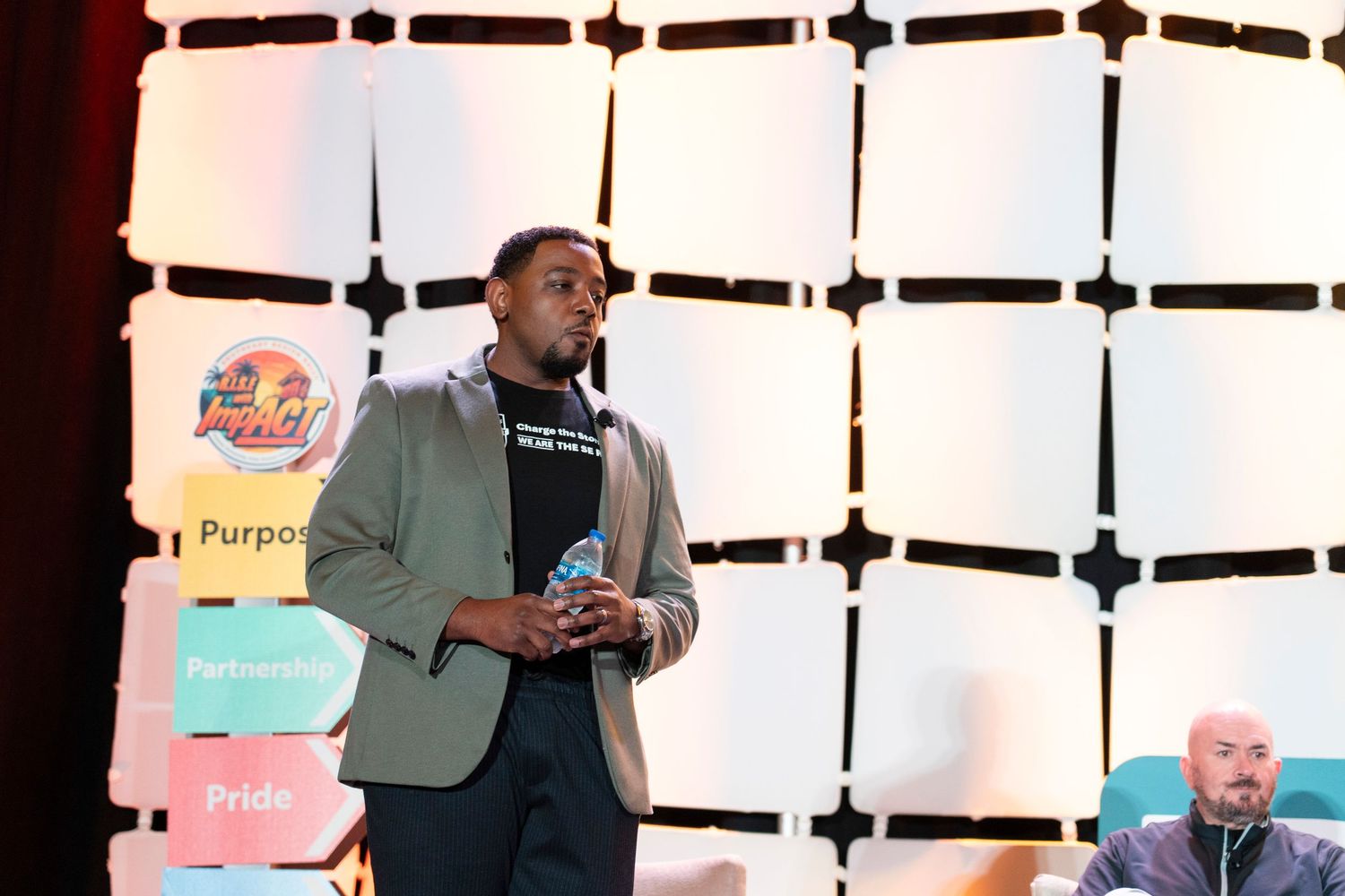 A man in a blazer speaks on stage near colorful Purpose, Partnership, and Pride signs at an event.