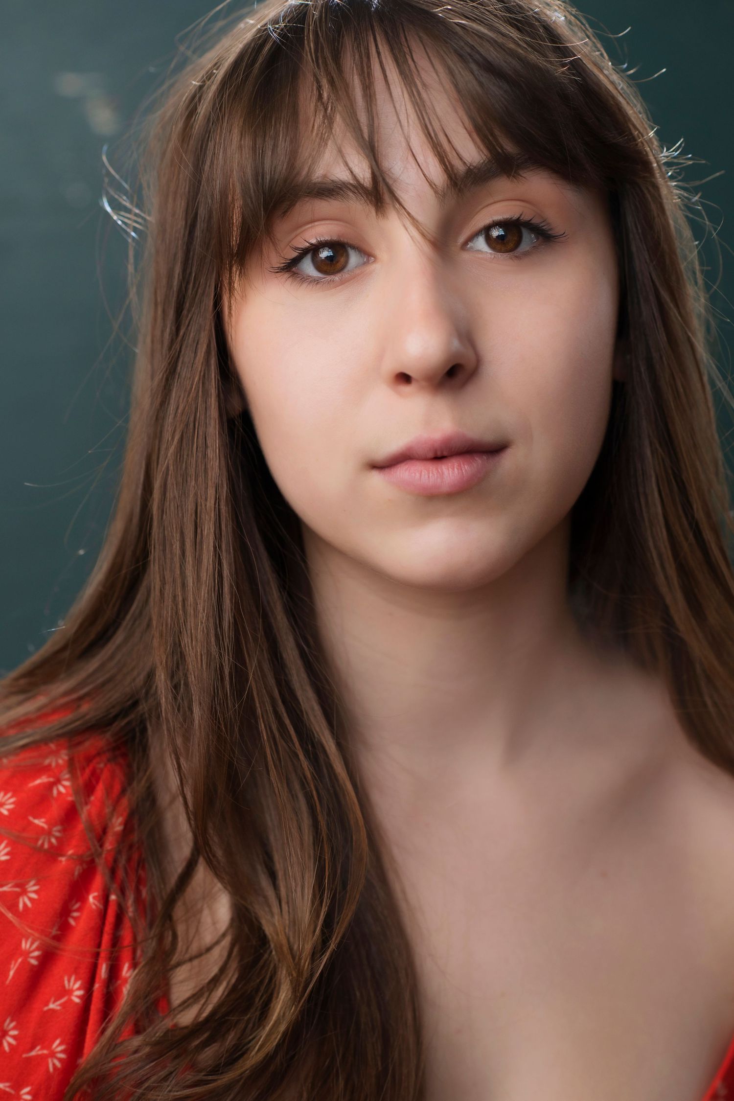 Close-up portrait of a young woman with long brown hair and bangs, wearing a red floral top against a teal background.