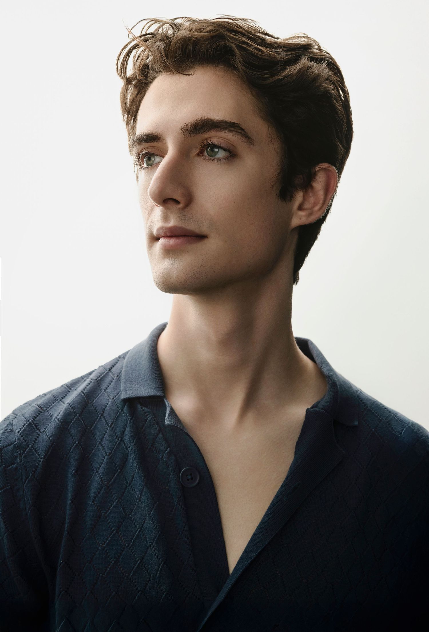 Handsome young man with green eyes and wavy hair in a navy diamond-pattern shirt on white background.