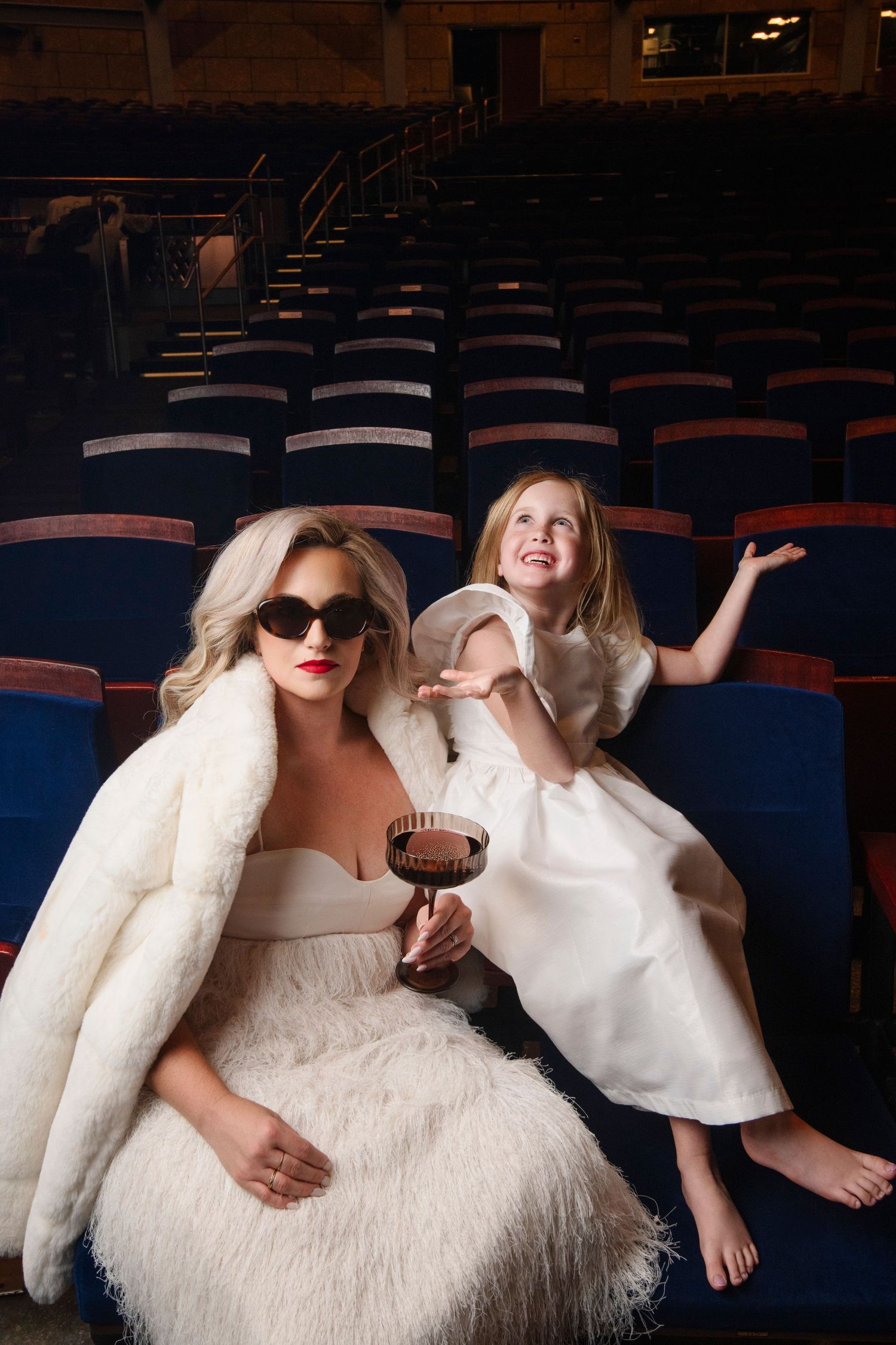 Woman in sunglasses and a white feathered gown holds a cocktail beside a laughing girl in a white dress in an empty theater.