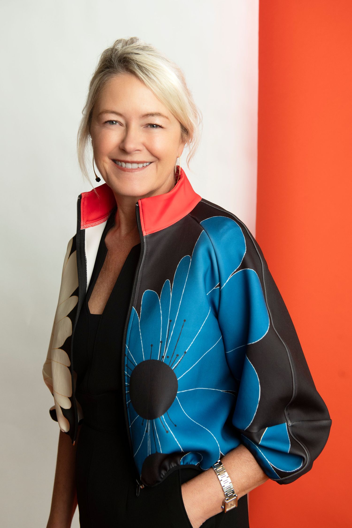 Smiling blonde woman in colorful floral jacket poses confidently against a white and orange background.