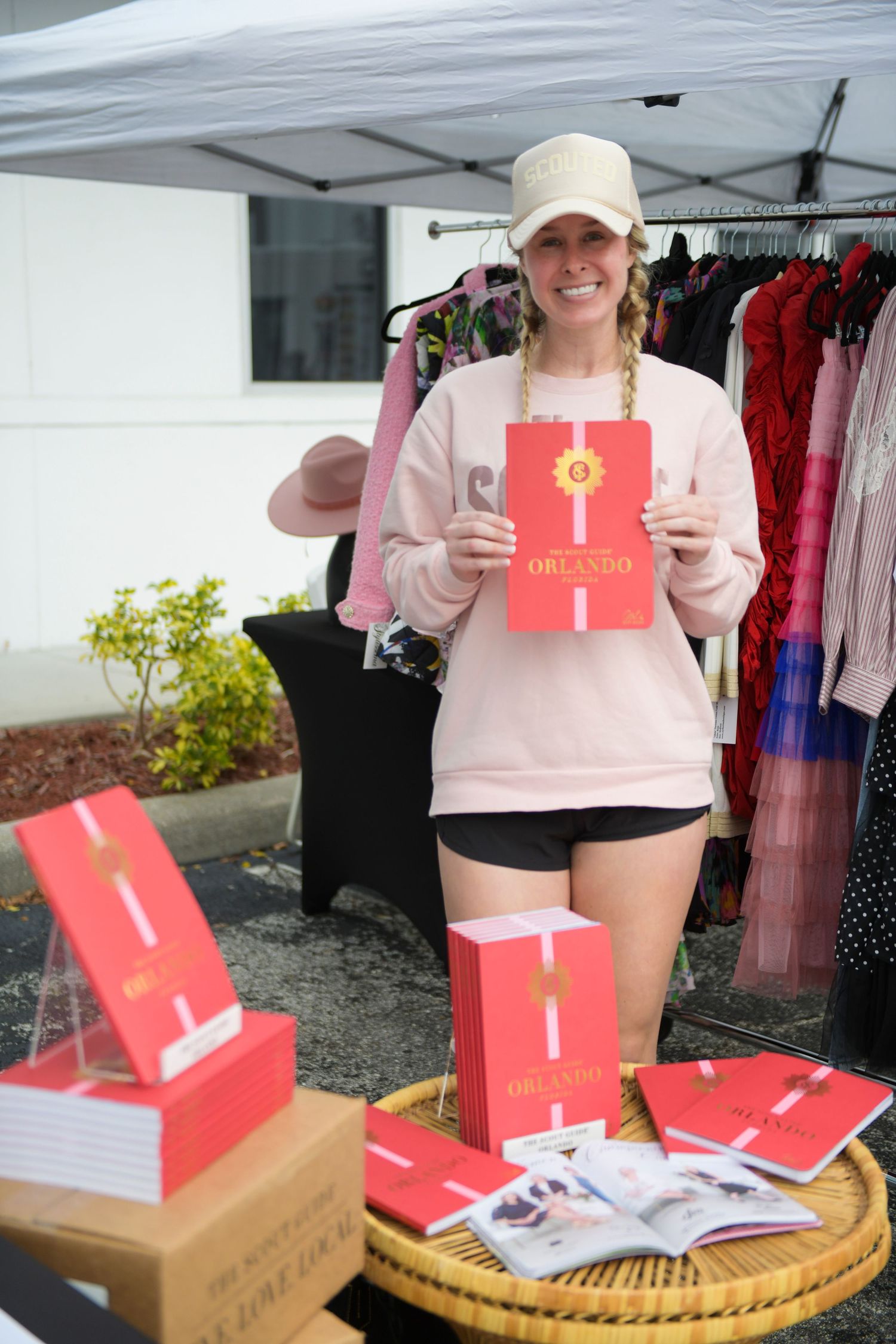 Woman in pink sweatshirt holding red Orlando book at outdoor market stall with books displayed on wicker table.